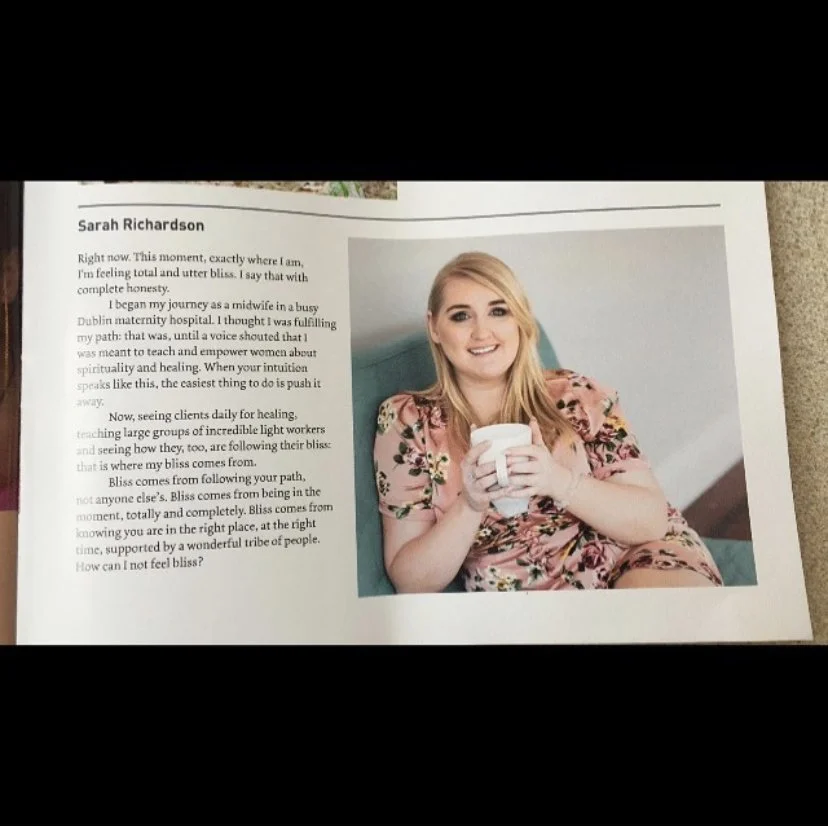 A woman with blonde hair wearing a pink floral dress, smiling, while holding a white mug and sitting on a teal-colored couch during the daytime.