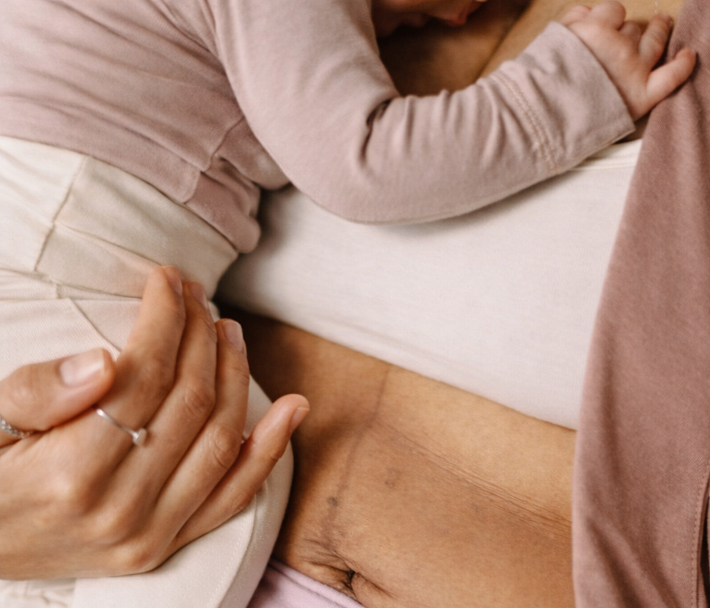 A person lying on their side in bed, exposing their lower abdomen and skin. The person's hand is gently resting on a child's back, who is lying beside them. The image focuses on the intimacy and comfort shared between them.