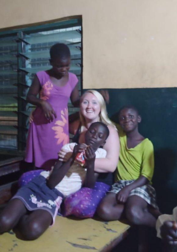 A woman with blonde hair surrounded by three African children inside a room with a window and yellow wall.