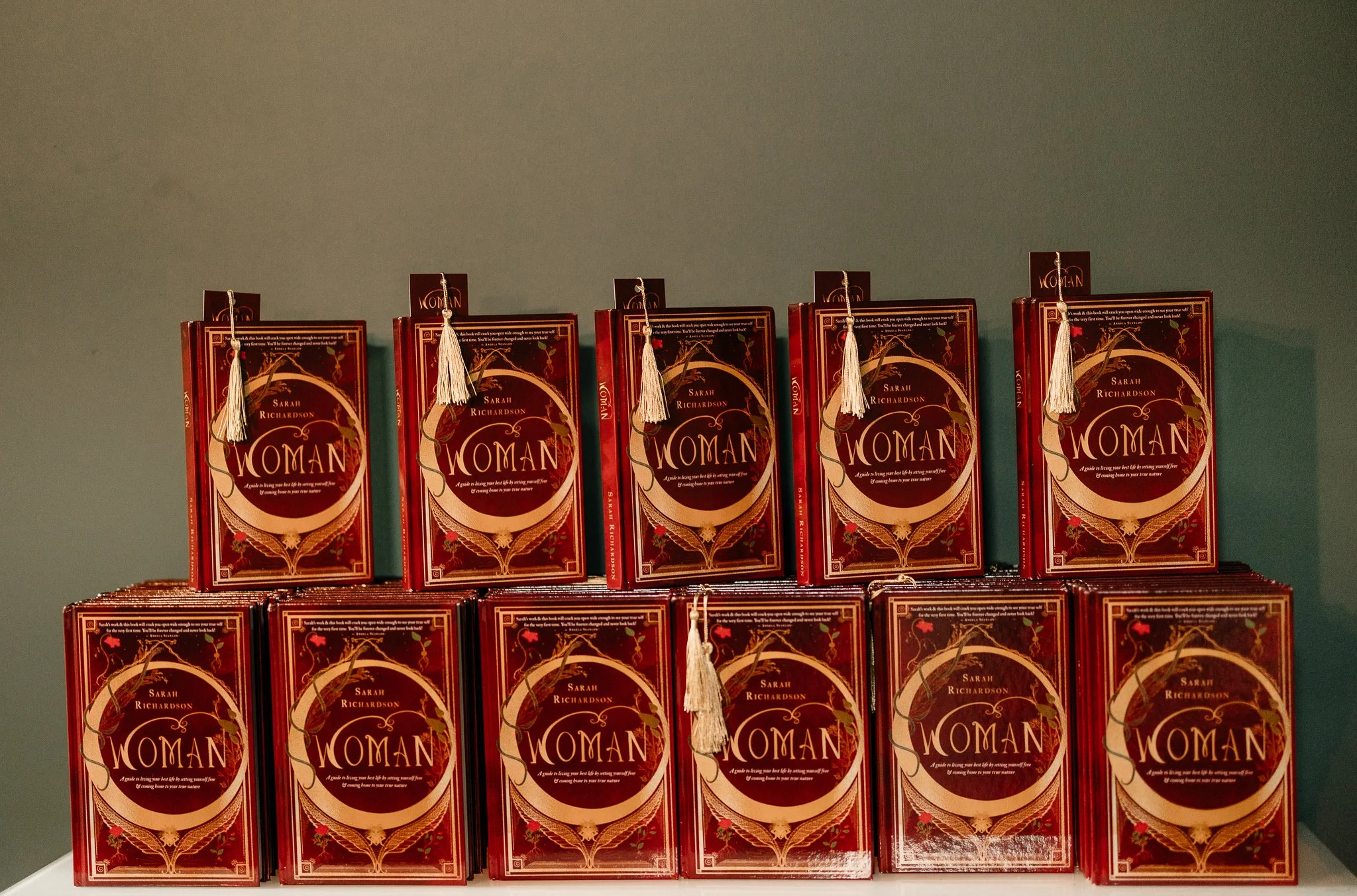 Stack of ten hardcover books titled 'Woman' by Sarah Richardson, arranged in two rows on a shelf with a plain green background.