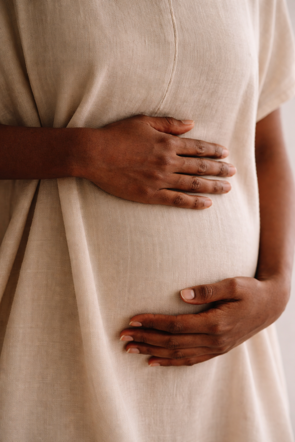 A person with dark skin gently cradling their pregnant belly with both hands, wearing a beige dress.