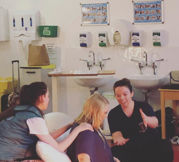 A woman talking and gesturing with two young girls in a pediatric clinic or hospital room with sinks, soap dispensers, and medical posters on the wall.