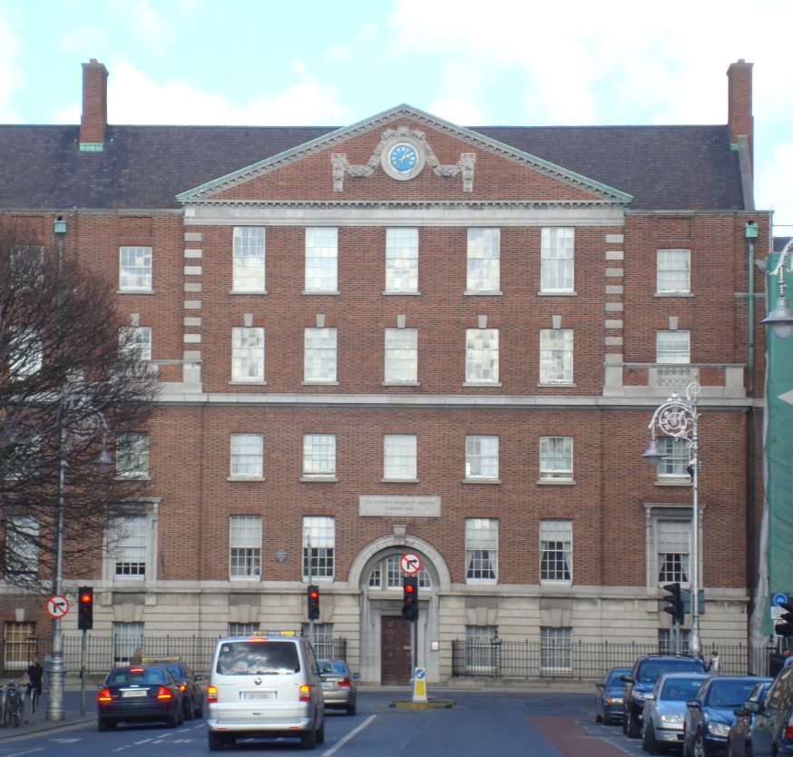 A large three-story brick building with a clock at the top, located on a city street with cars and traffic lights.