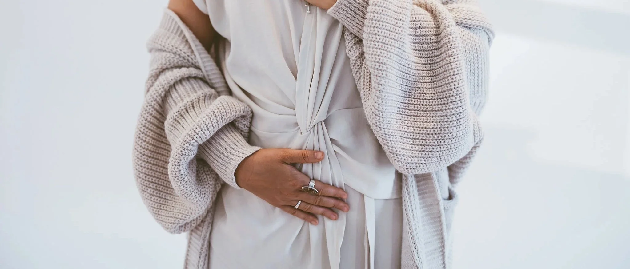 Close-up of a person holding their stomach, indicating possible stomach pain or discomfort, wearing a cozy beige knitted cardigan over a white outfit.