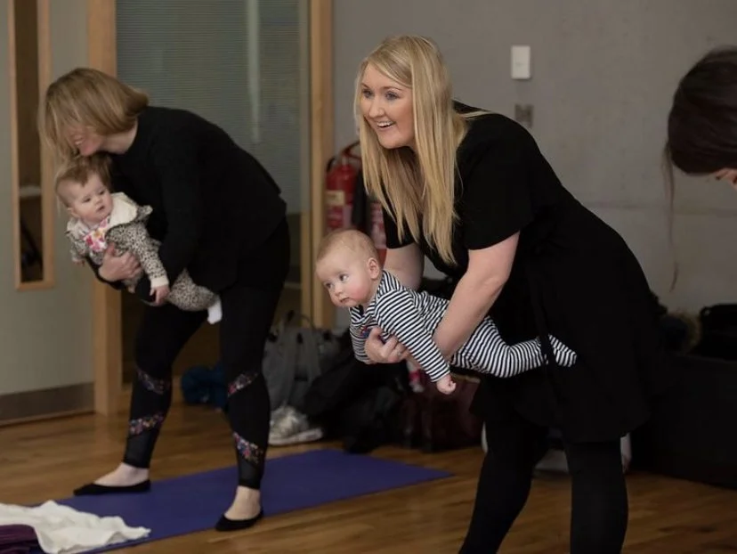 Two women are holding babies, one in a striped outfit and one in a leopard print, during a baby play session or class in a room with wooden floors and a blue mat.