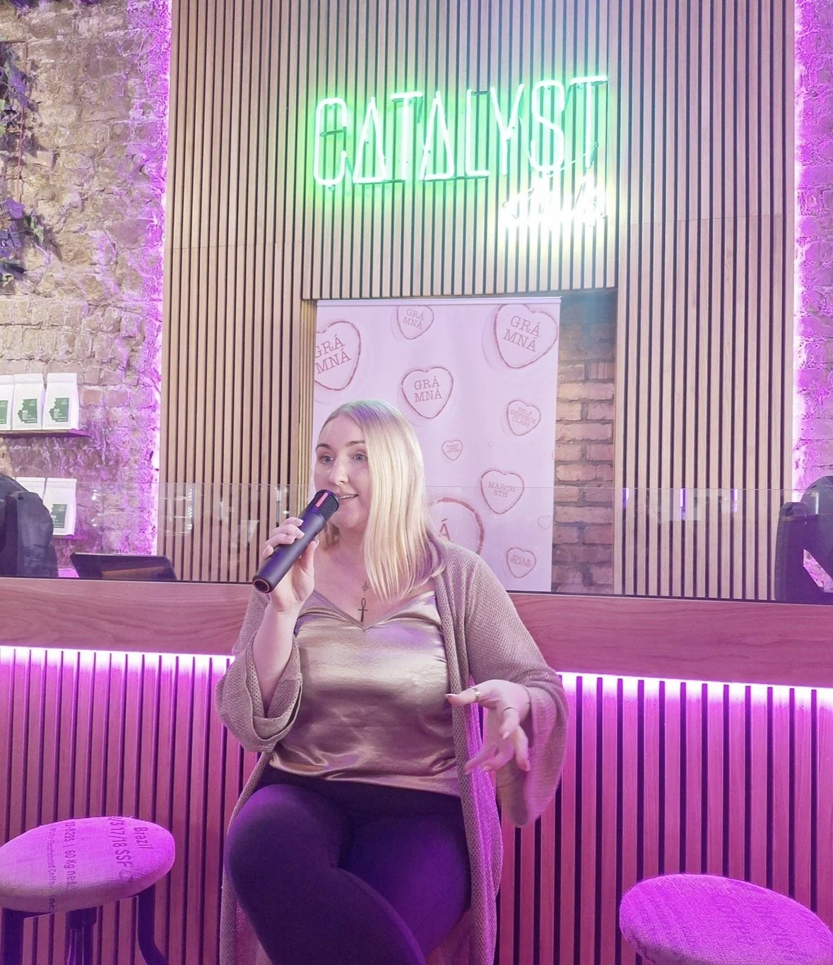 A woman holding a microphone, speaking at a cafe or restaurant with a purple and pink lit background, neon sign that says 'CATCHY YaST', a privacy shield, and a promotional poster in the background.