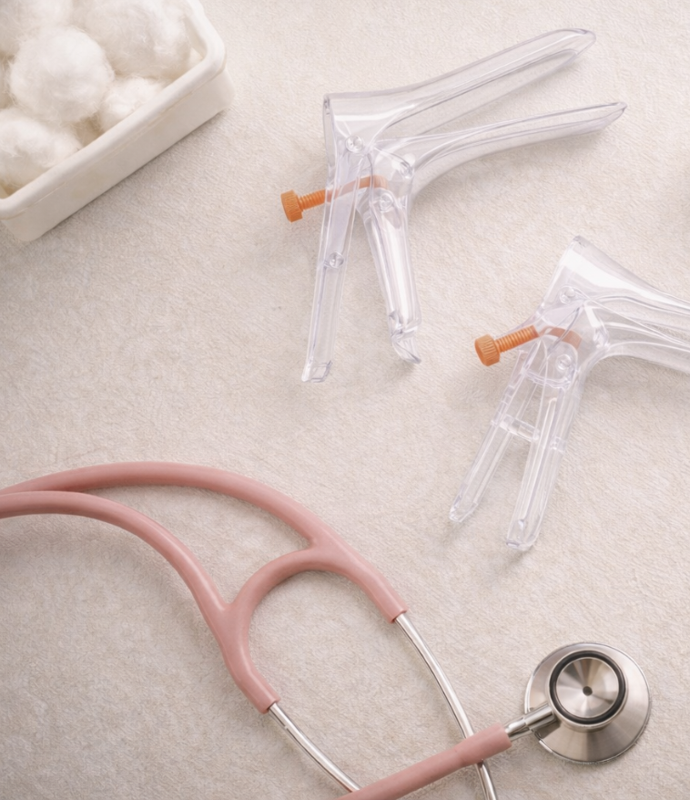 Medical tools including a pink stethoscope and two clear sprayers with orange knobs on a beige surface, with a white tray of cotton balls in the background.