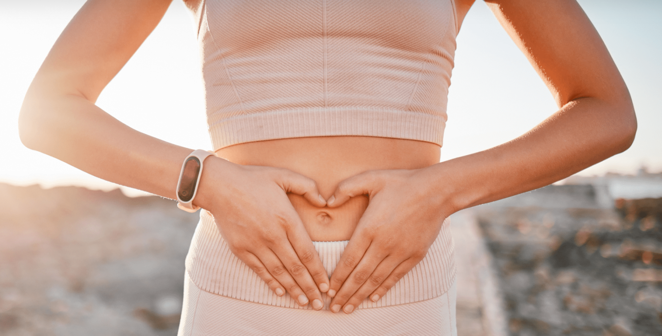 Person standing outdoors at sunset, making a heart shape with hands on stomach.