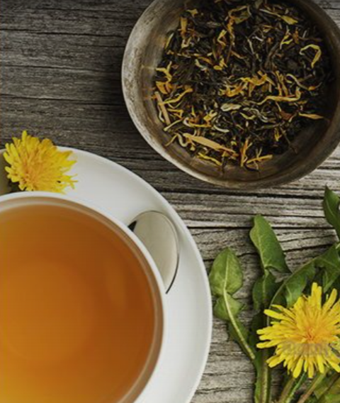 A cup of tea on a saucer with a teaspoon, a basket of loose herbal tea leaves, yellow flowers, and green leaves on a wooden surface.