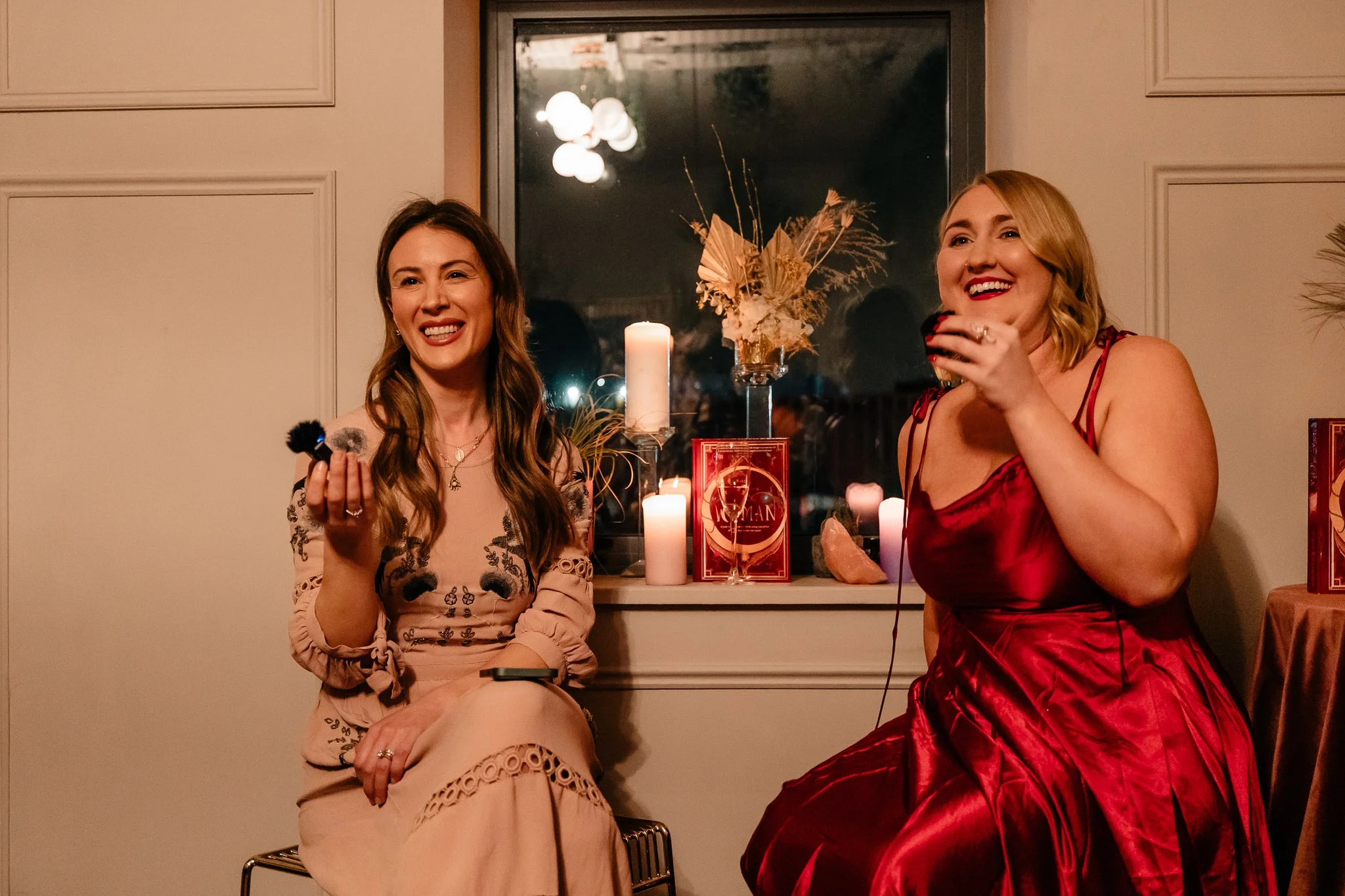 Two women sitting indoors at a cozy gathering, smiling and laughing, with candles and decorative items on the windowsill behind them.