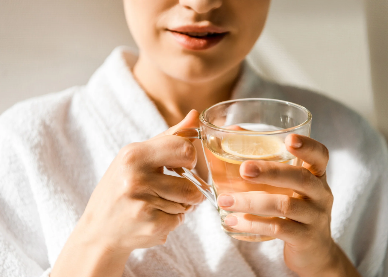 A woman in a white robe holding a glass cup of hot water with lemon slices inside.