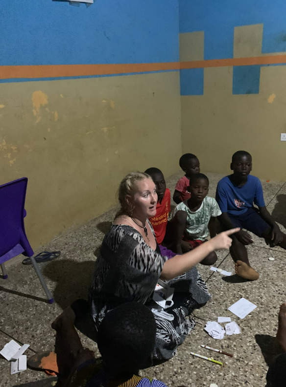 A woman with blonde hair seated on the floor, pointing to her right, surrounded by young children in a room with painted blue and yellow walls and a purple chair nearby.