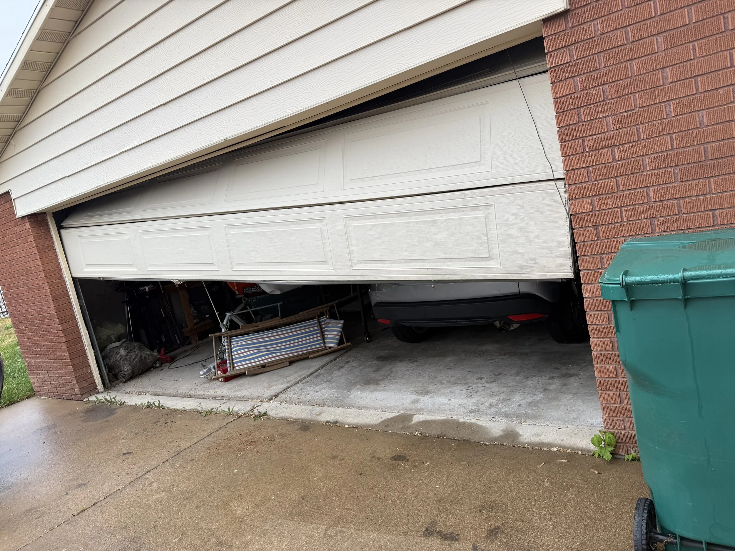 The image shows an open garage with a white garage door, a brick pillar on the right, and various items stored inside, including a striped chair, a boat, and bags. A green trash bin is outside on the right.