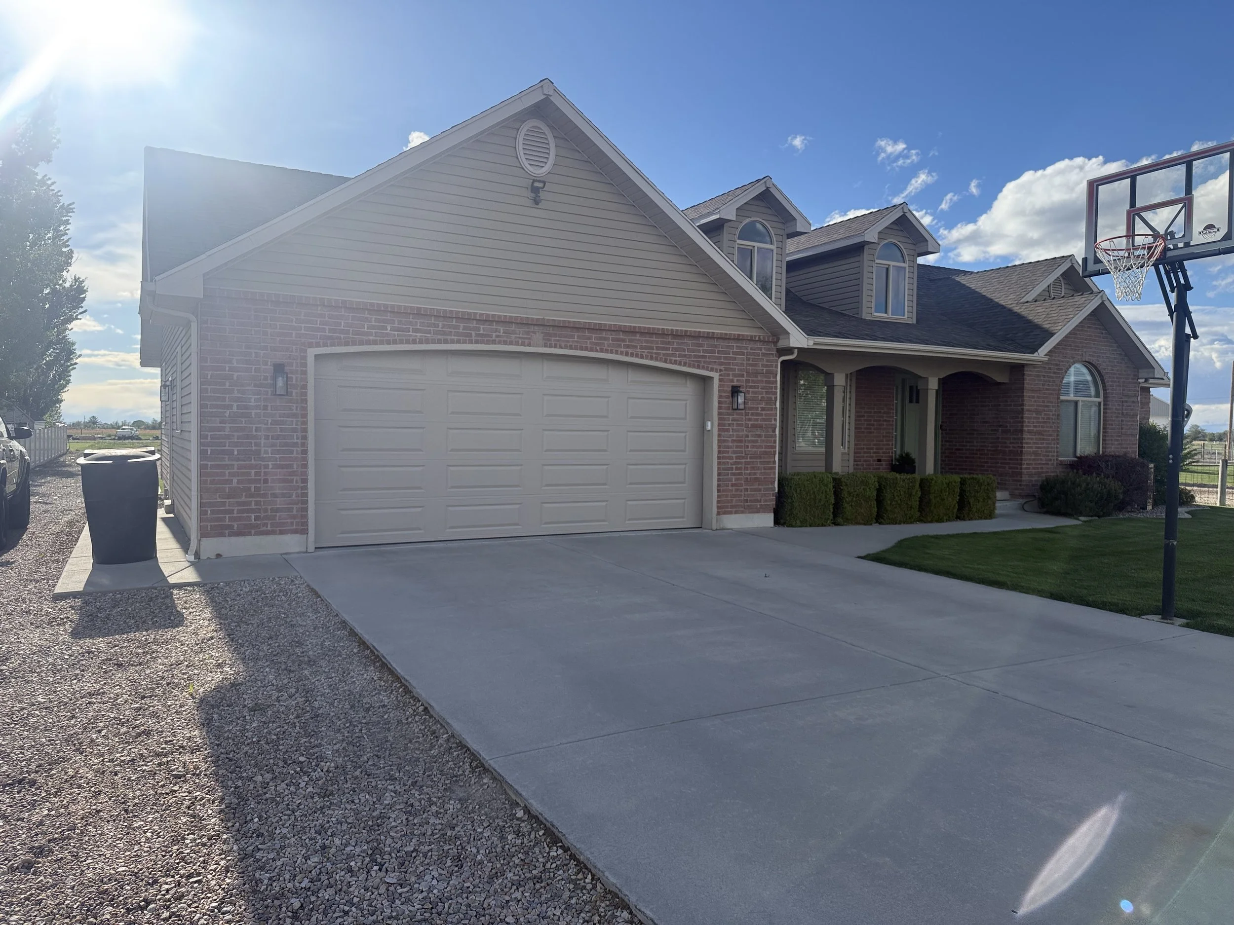 A suburban house with brick and siding exterior, a white garage door, and a basketball hoop outside on a sunny day with a blue sky and some clouds.