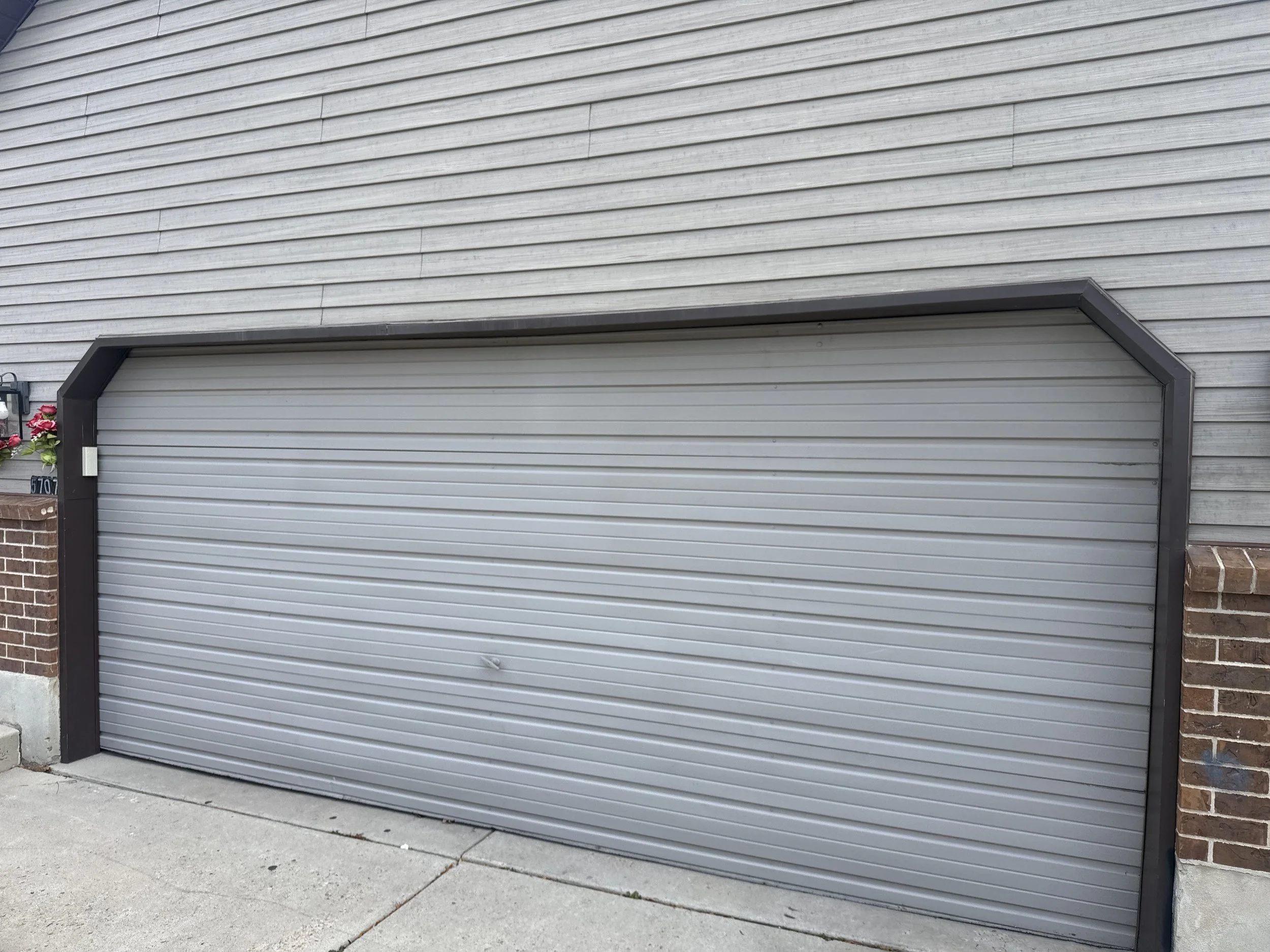 Gray metal garage door with horizontal ridges, bordered by brick walls on each side, situated on a concrete driveway.