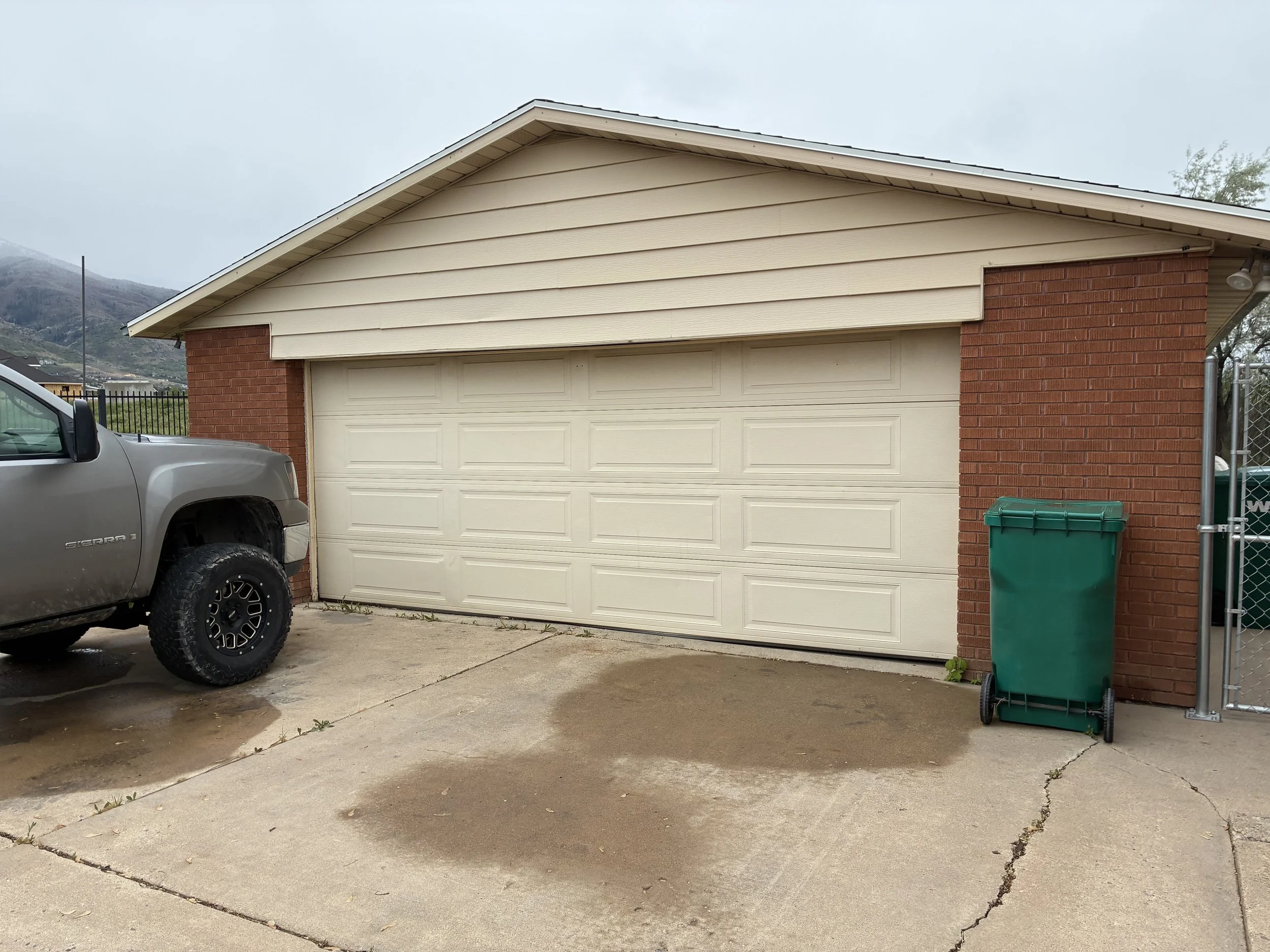 A suburban house with a beige garage door, red brick walls, and the roof shining under an overcast sky. A silver pickup truck is parked partly in front of the garage, and a green trash bin is beside the house near a chain-link fence. A mountain and some trees are visible in the background.