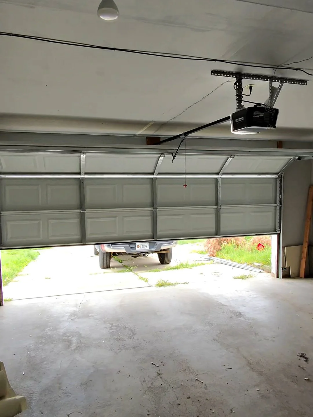 Empty garage with an open garage door, showing a parked vehicle outside. The garage has concrete flooring and a ceiling with overhead lighting and an automatic door opener.