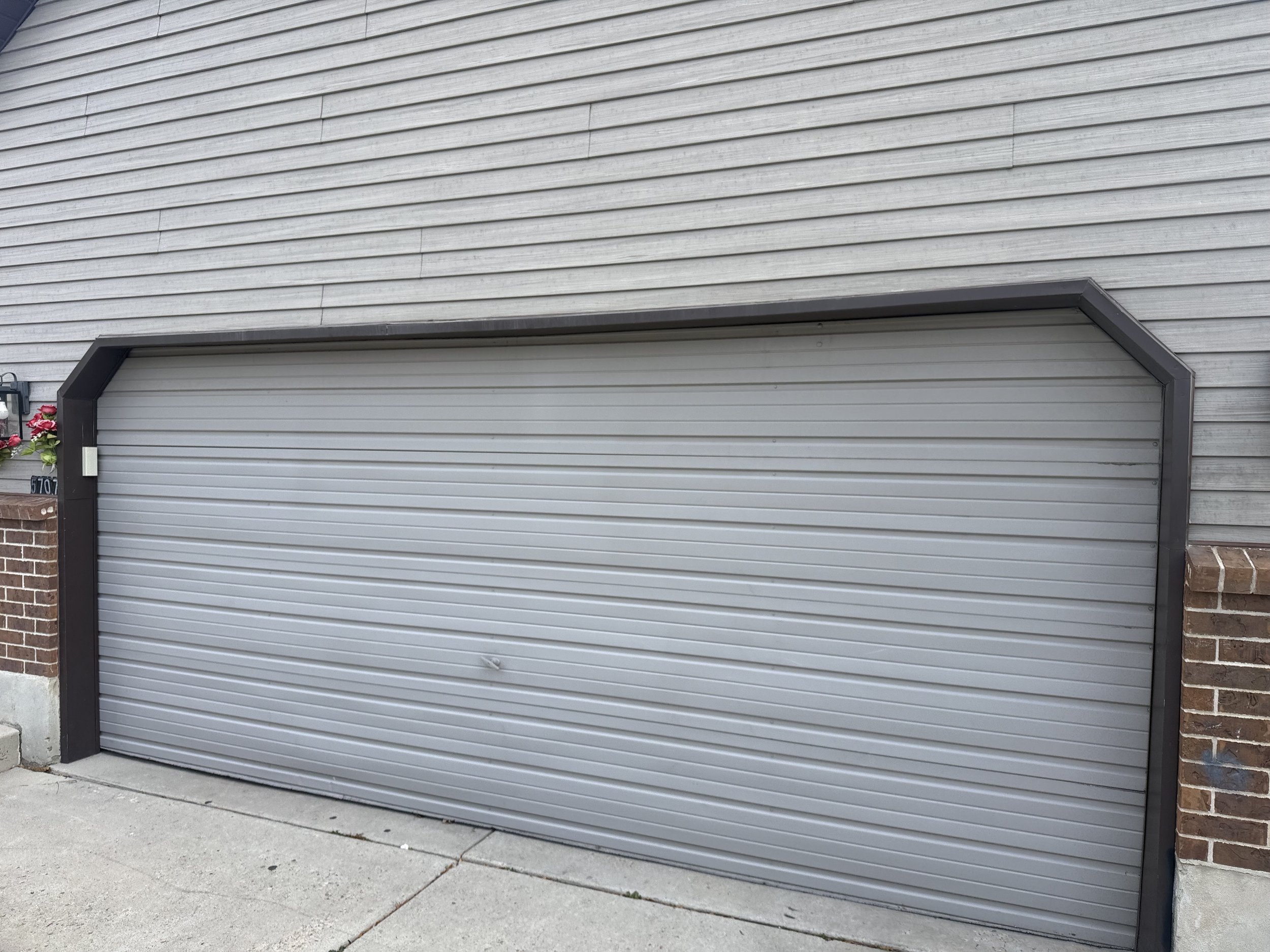 A gray garage door with horizontal lines, frame, and brick wall on the right side, with a patch of sidewalk and pavement in front.
