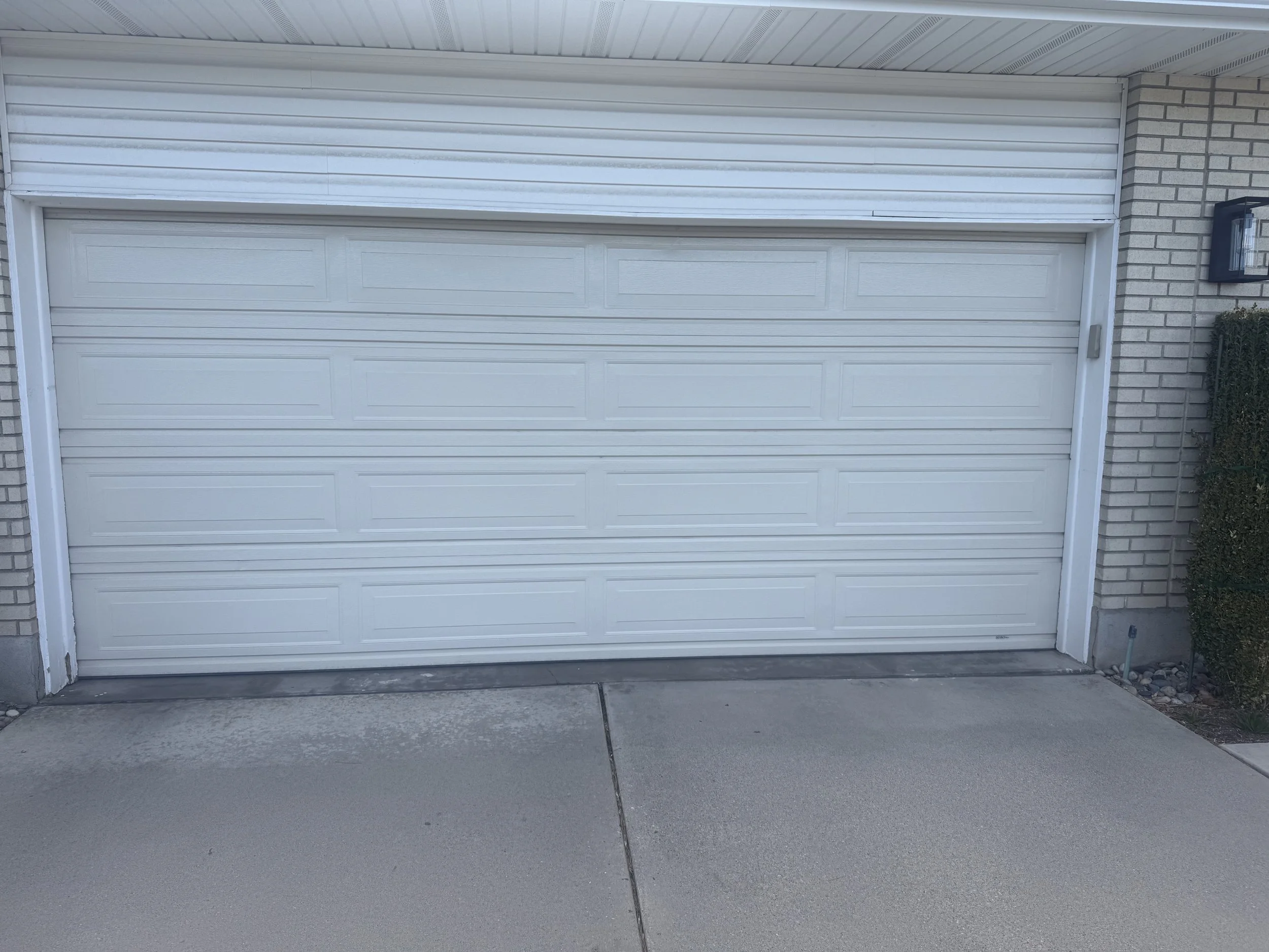A white garage door framed by brick walls on both sides, with a black outdoor wall lantern on the right side and some green shrubbery near the bottom right corner. The concrete driveway in front has visible cracks.