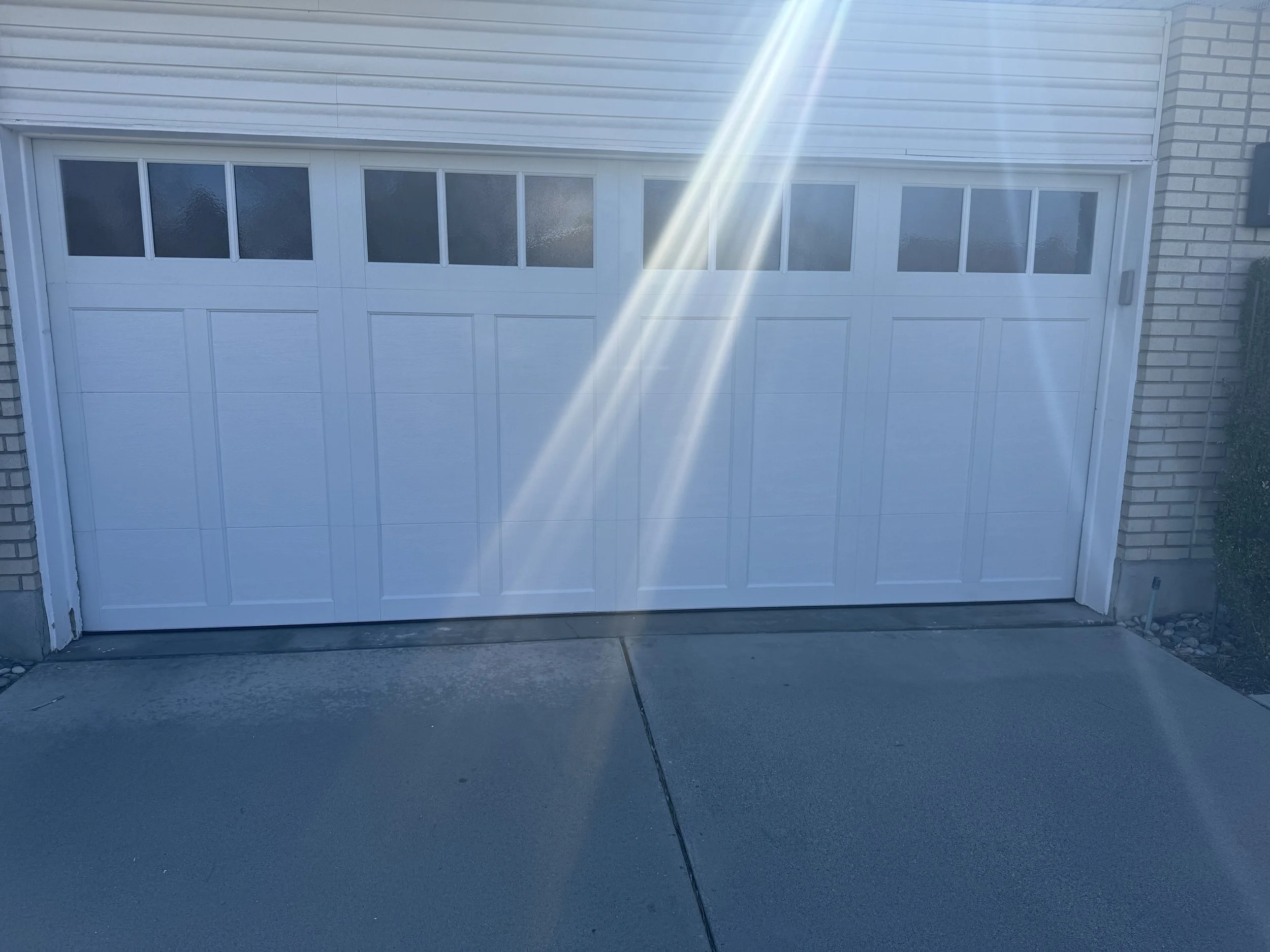 A white garage door on a brick house. Sunlight with lens flare appears at the top of the door. The driveway is concrete, with a black hose extending across it.
