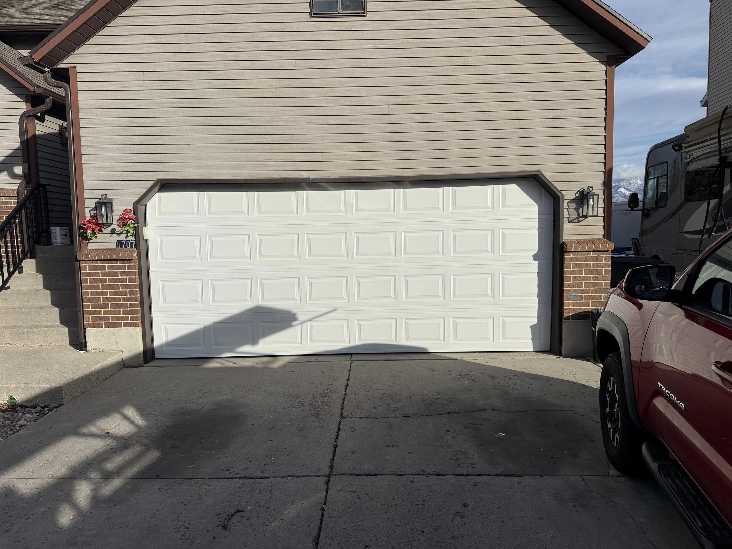 Front of a house with beige siding, a white garage door, brick steps, and pink flowers in black planters on either side of the steps. There is a silver vehicle parked on the driveway to the right.