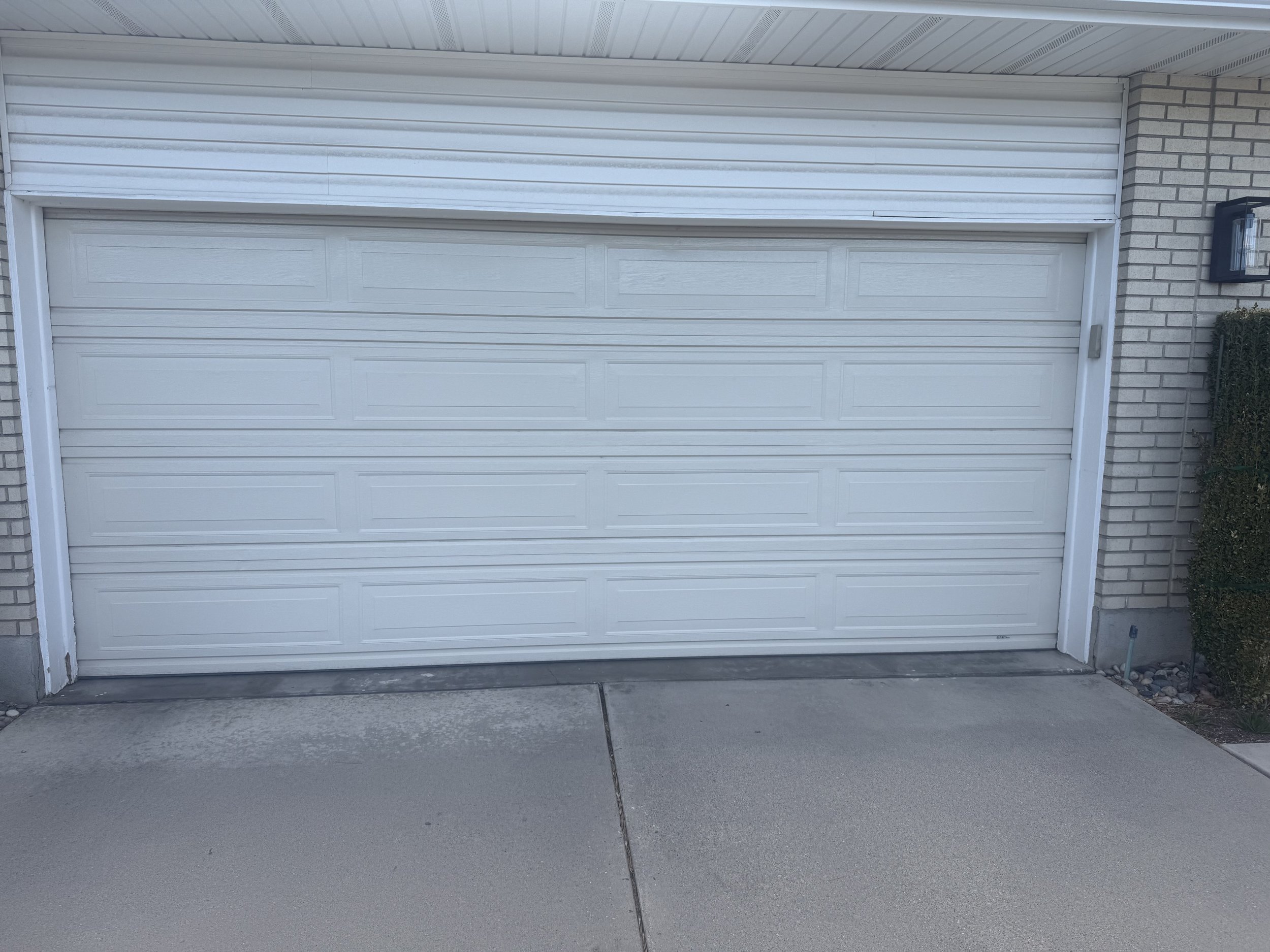 A closed white garage door with a brick house on the right side, a black outdoor wall light on the right, and some plants at the bottom right. The driveway in front is made of concrete.