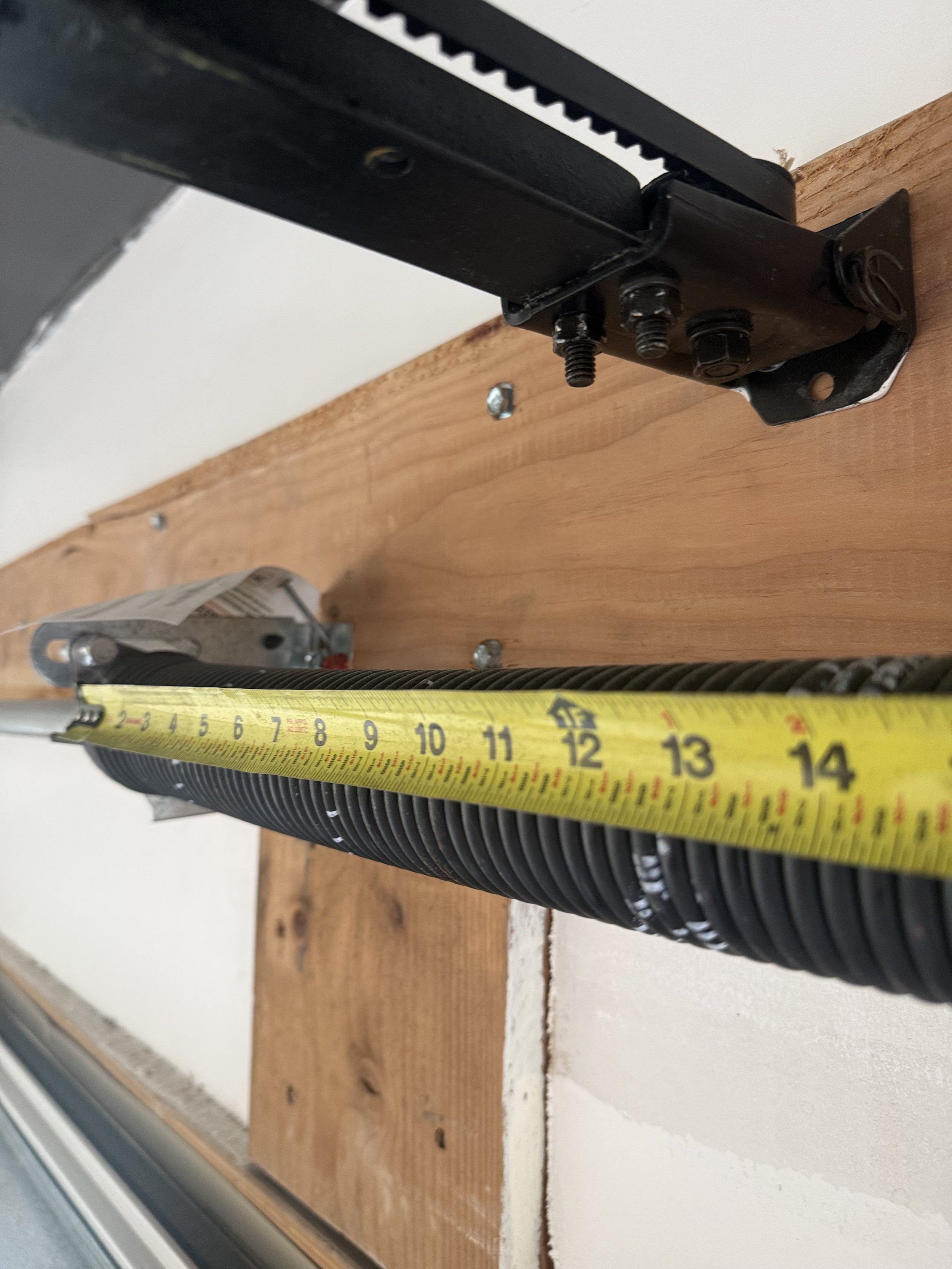 Close-up of a black metal hinge or mechanical part attached to a wood surface, with a measuring tape stretched out beneath it.