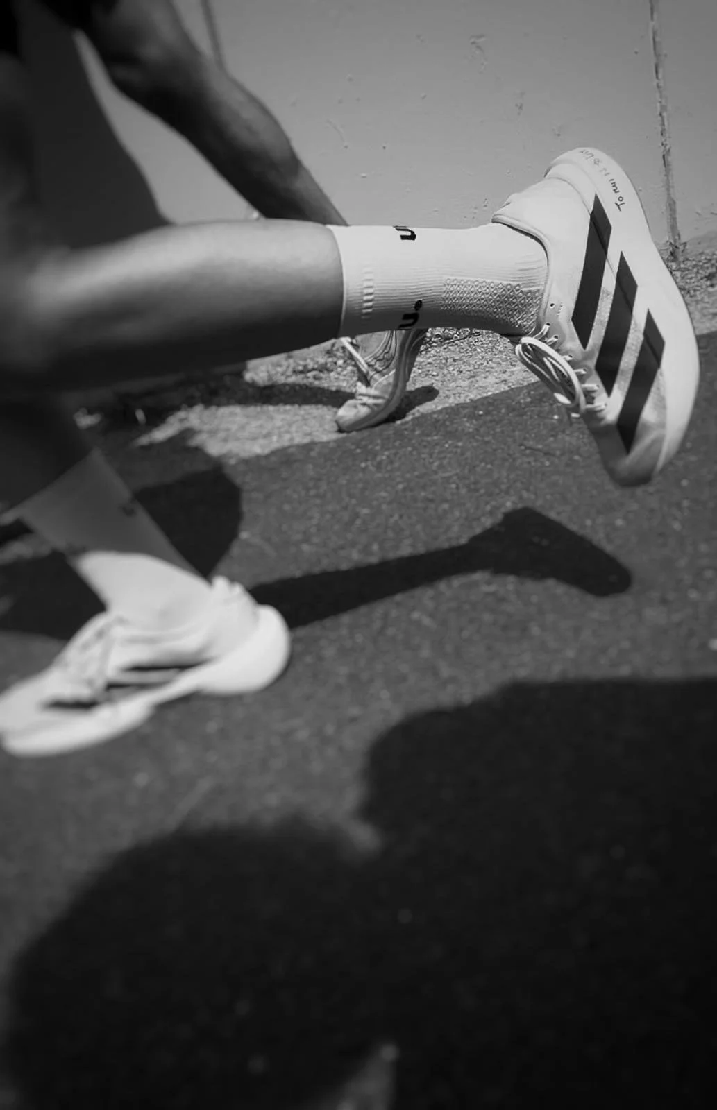 A runner wearing athletic shoes, socks, and shorts bent over on a track or pavement, with a wall in the background and shadow cast on the ground.