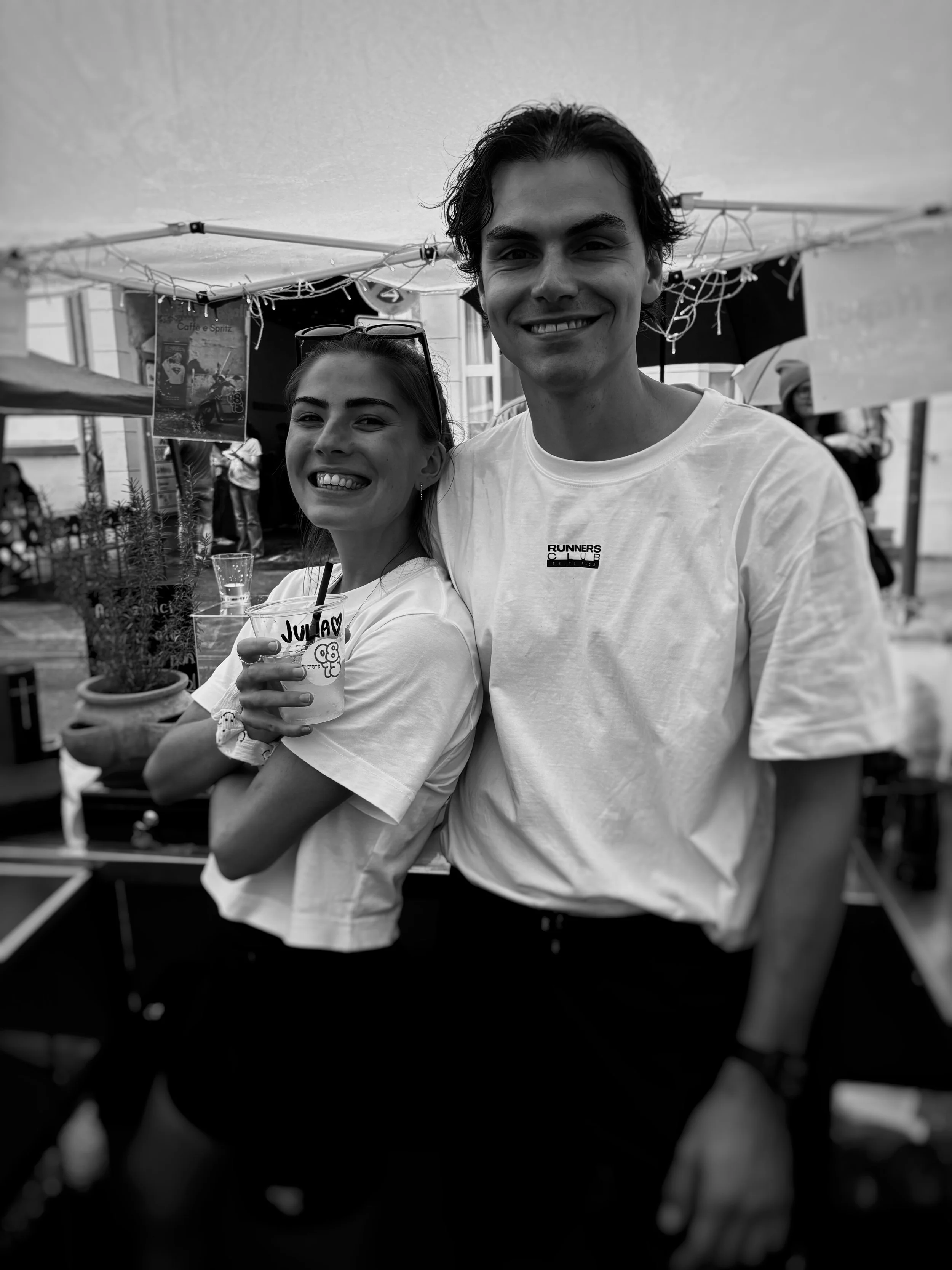 Two people smiling, a young woman and a young man, standing close together outdoors at a festive event or market.