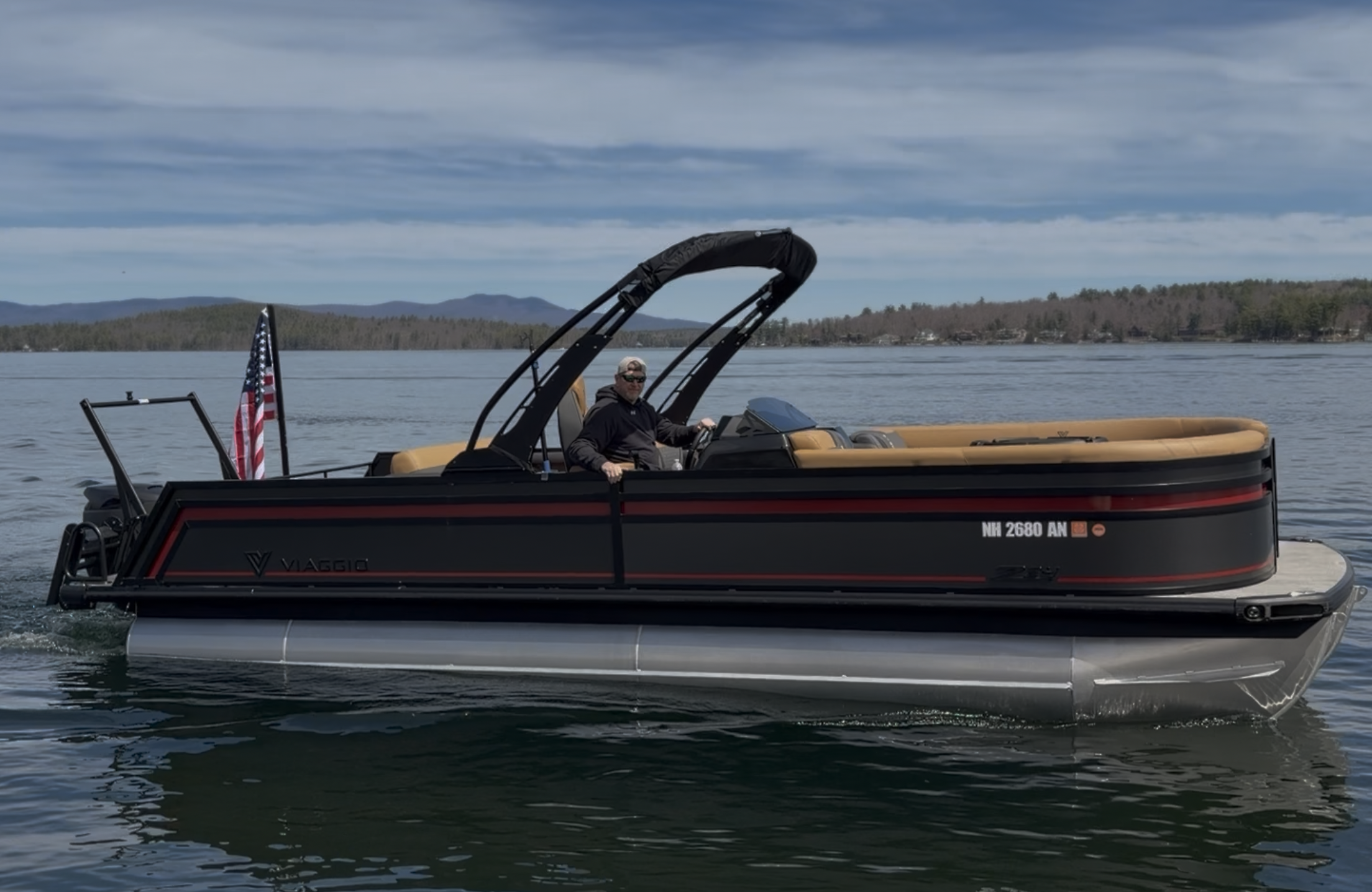A man wearing sunglasses and a dark jacket is driving a black and tan motorboat on a lake, with a background of distant trees and mountains under a partly cloudy sky.
