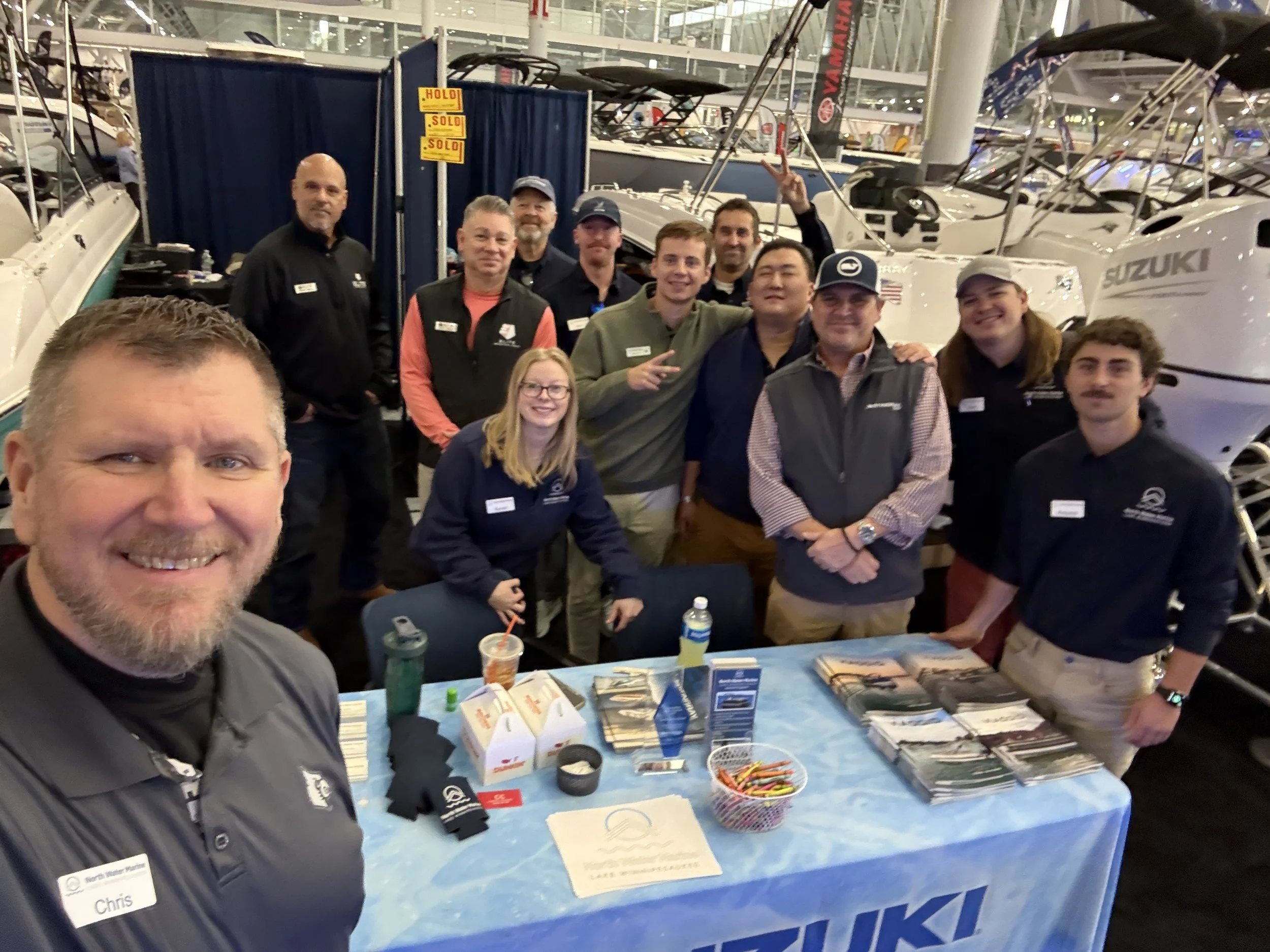 Group of people at a boat exhibition behind a table with brochures and promotional items, surrounded by boats in an indoor show.