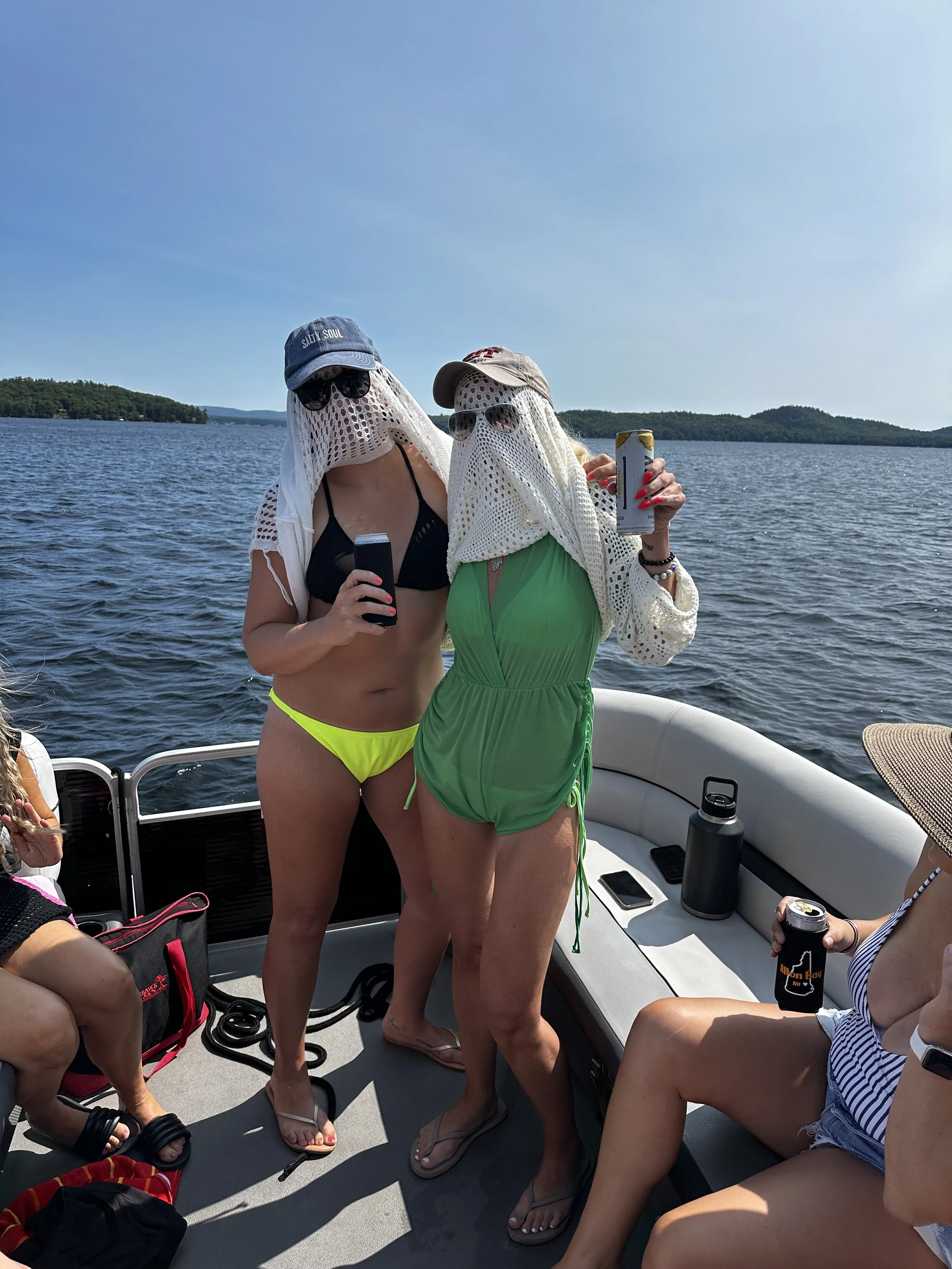 Group of women on a boat near a lake, wearing beach attire, hats, and sunglasses, holding drinks, with trees and hills in the background.