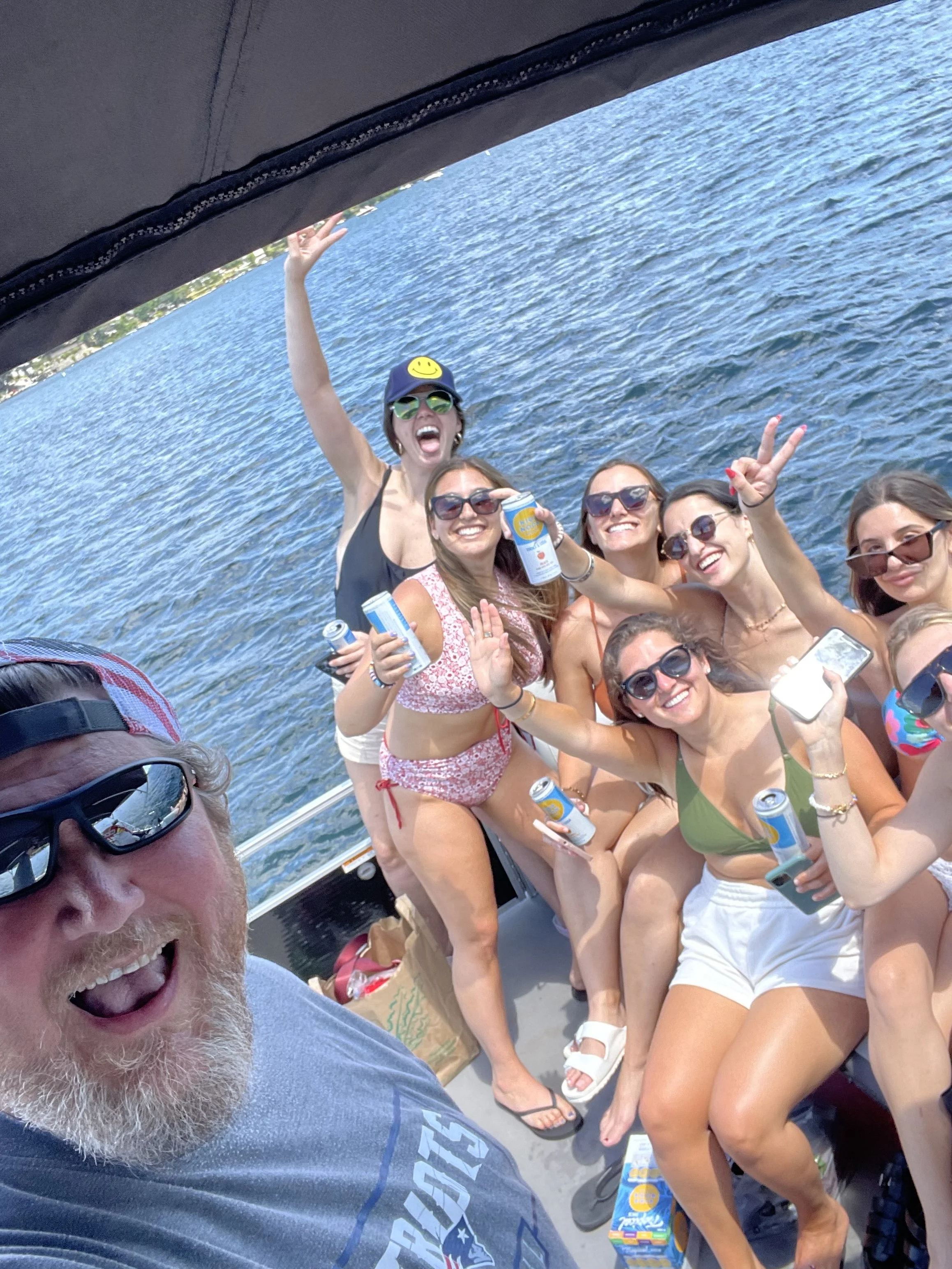 Group of women and one man on a boat, smiling and enjoying a sunny day by the water, some holding drinks, some making peace signs, with a scenic water view in the background.