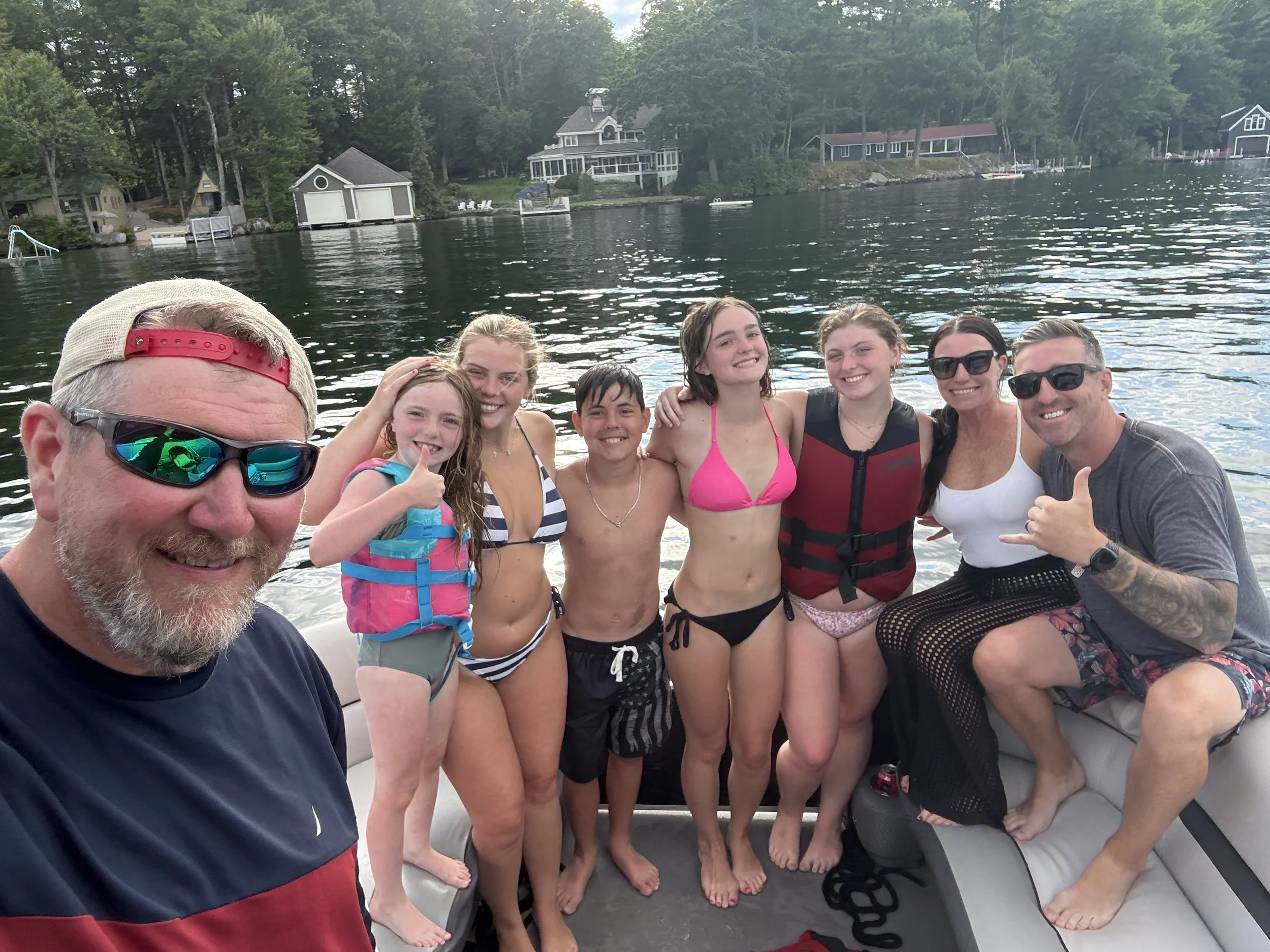 A group of eight people on a boat by a lake, with several houses and trees in the background. Some people are wearing swimsuits and sunglasses, smiling and posing for the photo.