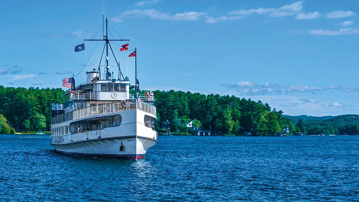 The M/S Mount Washington cruise ship navigating the deep blue waters of Lake Winnipesaukee on a clear day, with a dense green shoreline and summer homes in the background.