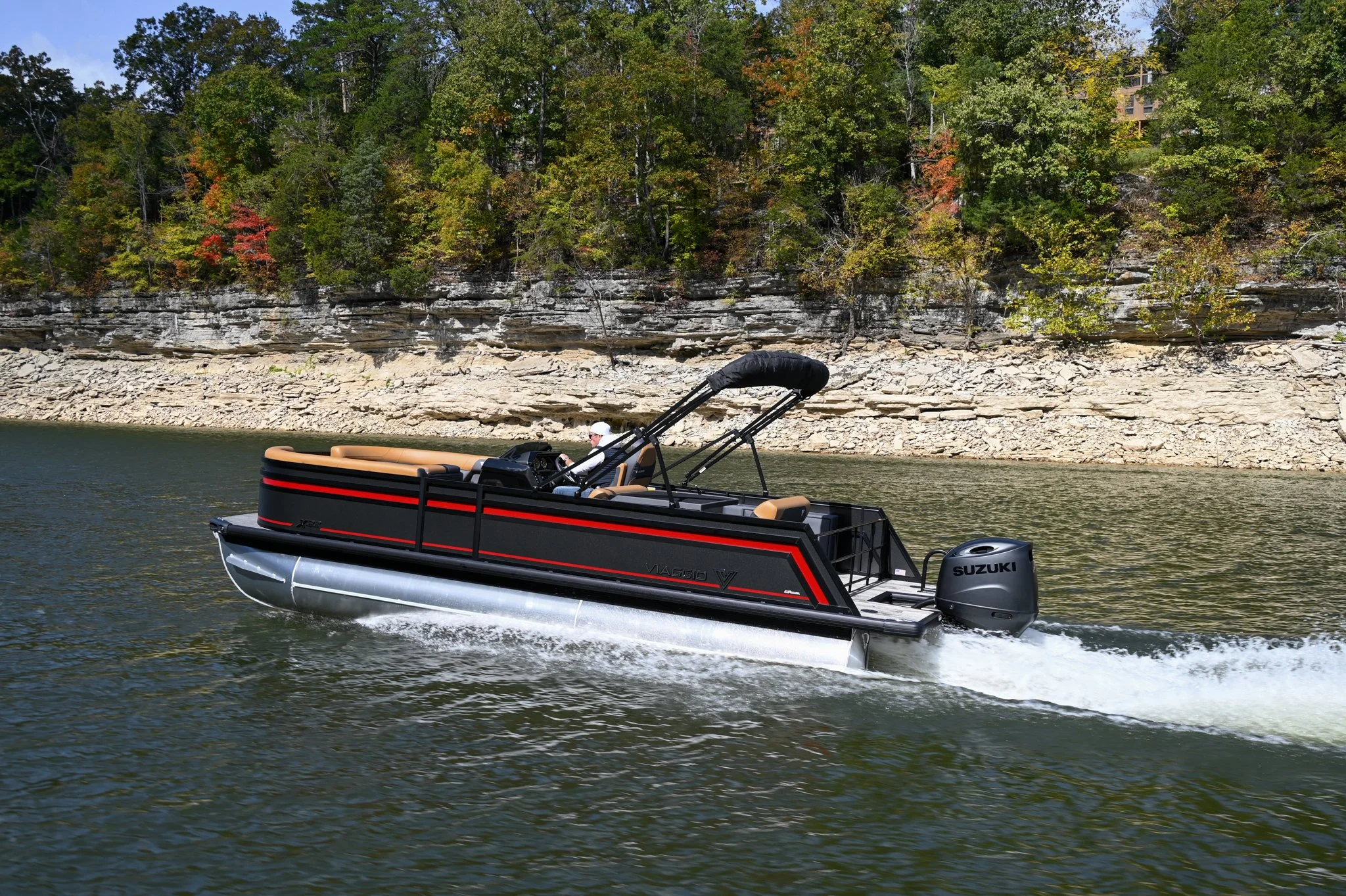 A black and red pontoon boat with tan seats cruising on a river, with a person in a white cap guiding it, and a background of rocky cliffs and trees with some fall foliage.