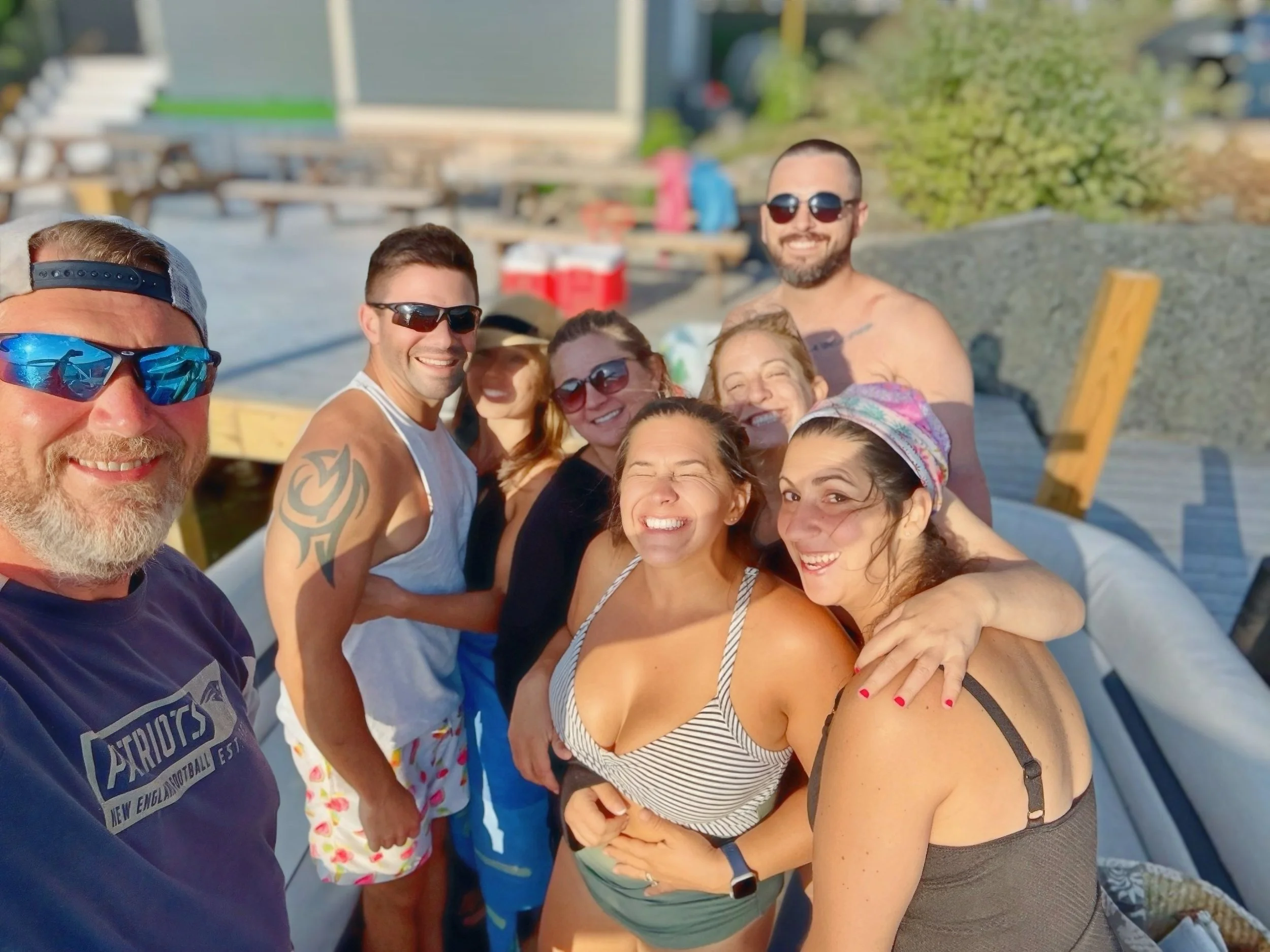 Group of smiling people taking a selfie outdoors on a sunny day, dressed in casual summer clothing, with a man in a purple shirt and sunglasses on the left and several women and men behind him.