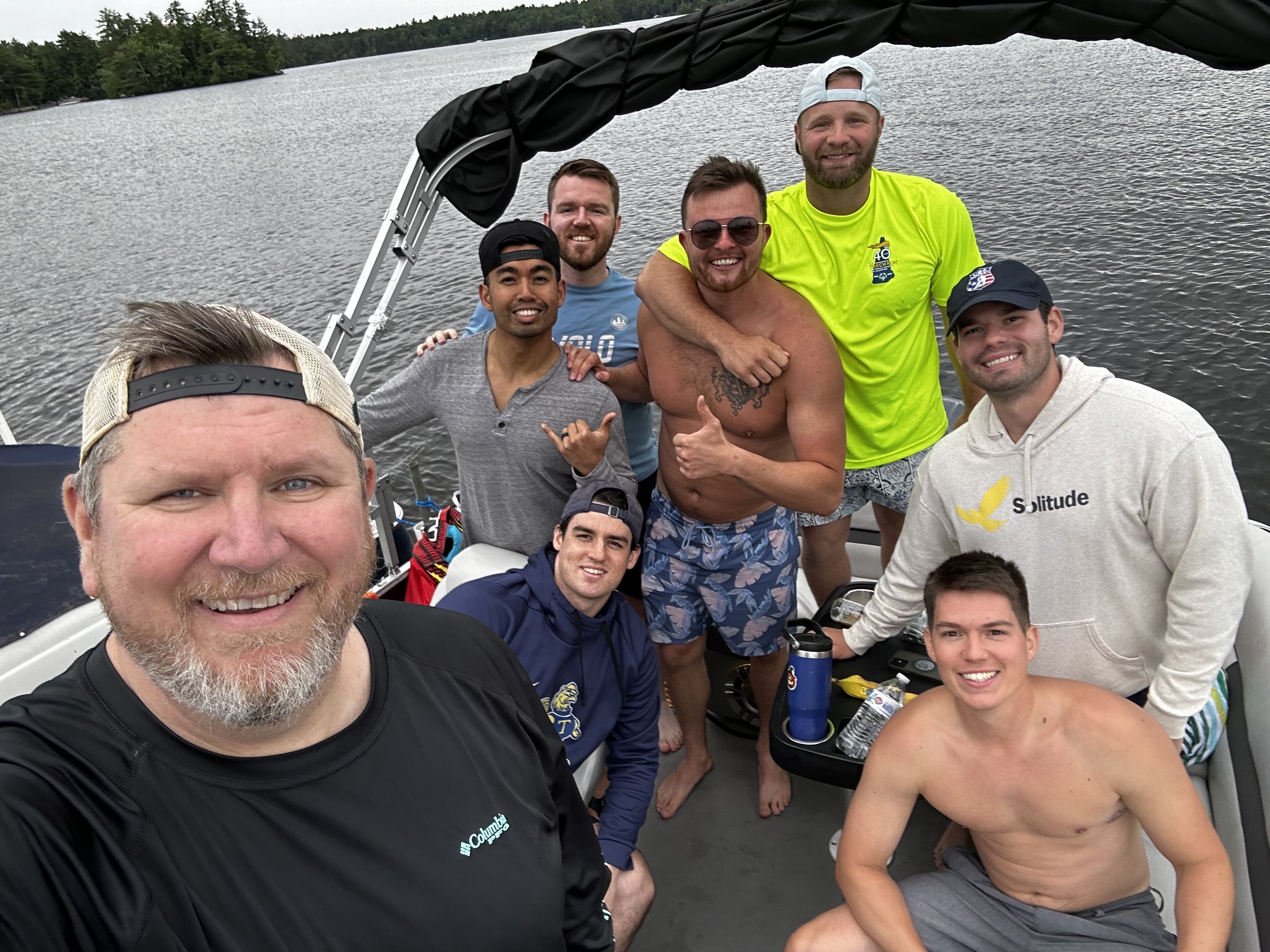 Group of six men on a boat, smiling and posing for a selfie near a lake with trees in the background. Some men are shirtless, and one is wearing sunglasses and a bright yellow shirt. There are water bottles and personal items visible on the boat.