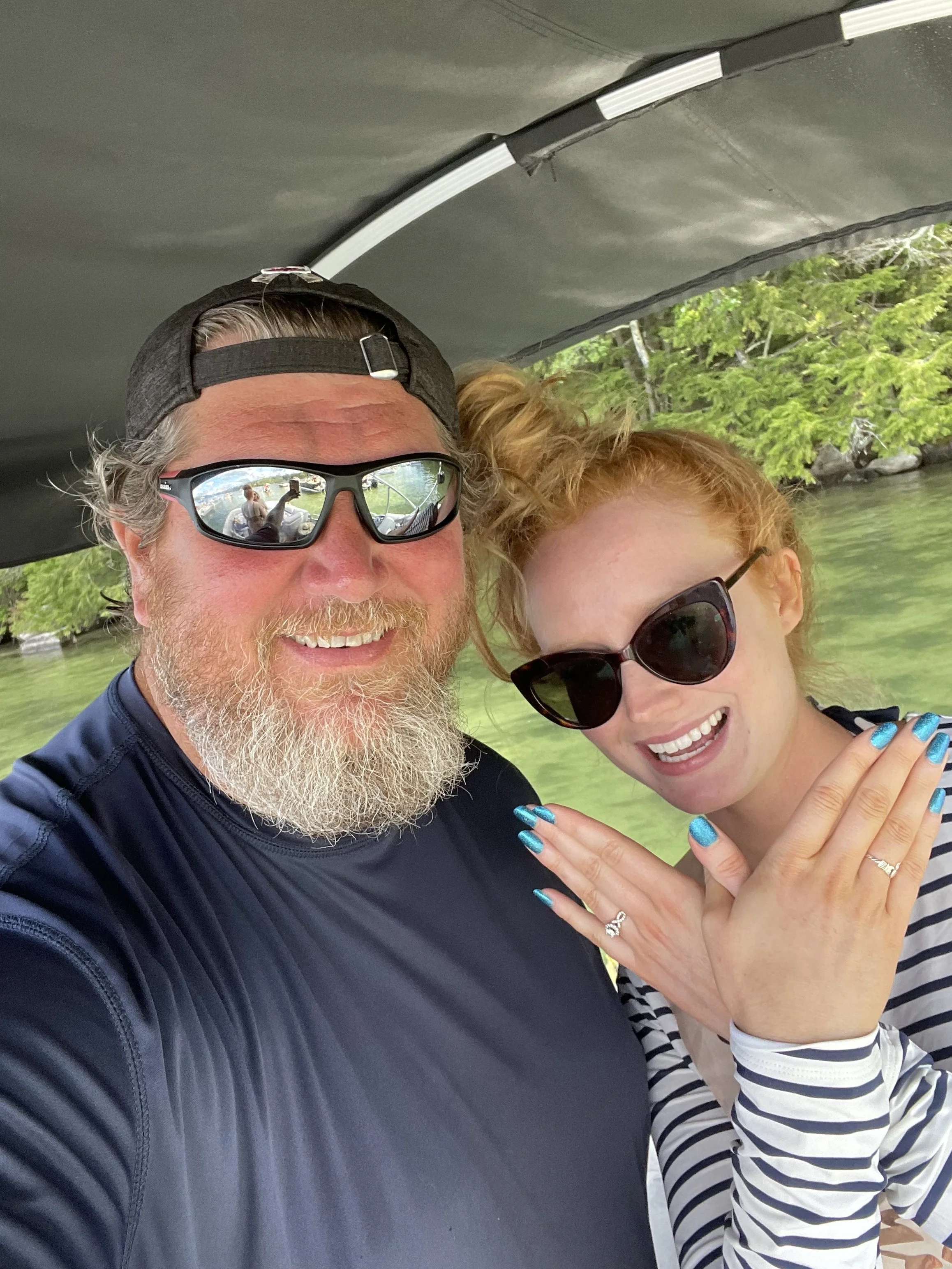 A man and woman smiling together on a boat, with water and greenery in the background. The man has a beard, sunglasses, and a black cap, and the woman has red hair, sunglasses, and painted nails.