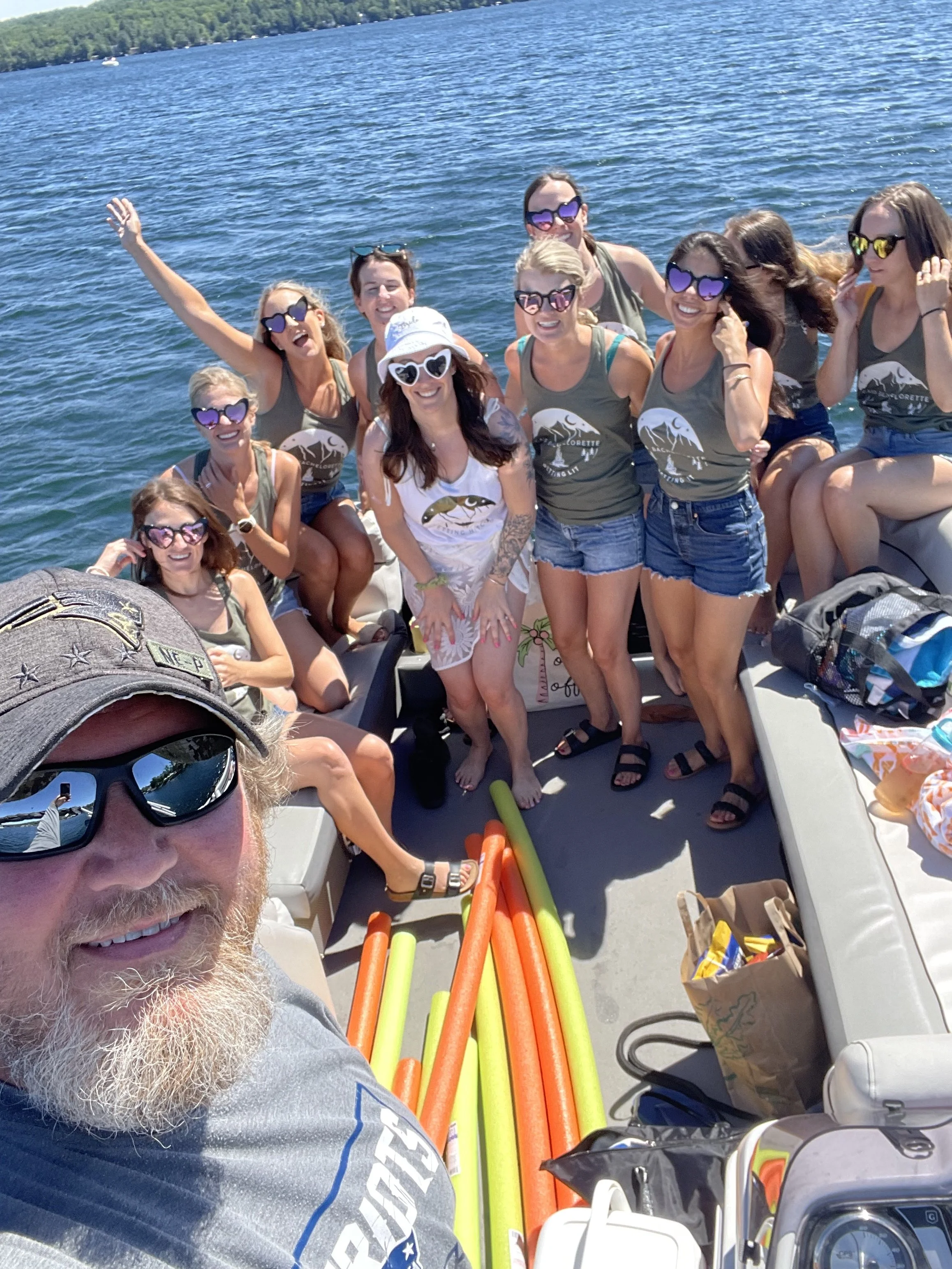 A group of smiling women and a man on a boat, wearing sunglasses and tank tops with a fish logo, enjoying a sunny day on a lake with trees in the background. There are colorful pool noodles on the boat and various bags and items around.