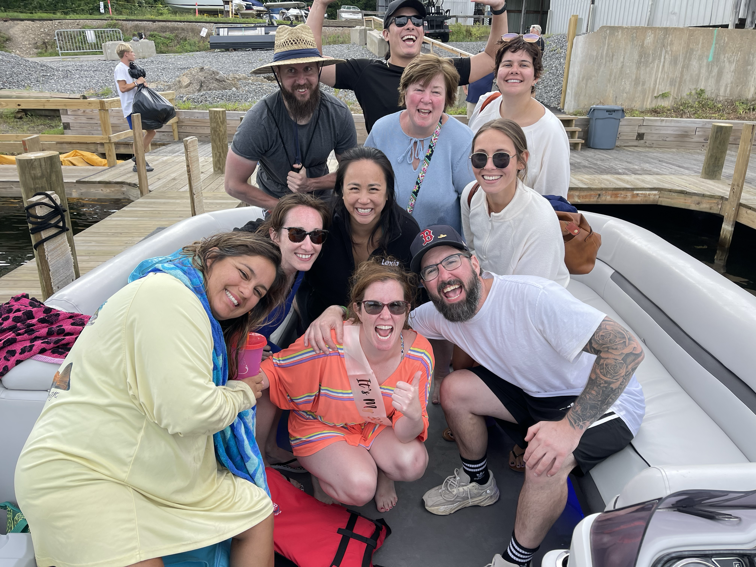 Group of smiling people on a boat, celebrating outdoors near a wooden dock, with a young boy in the background holding a bag, and a man making a joyful gesture.