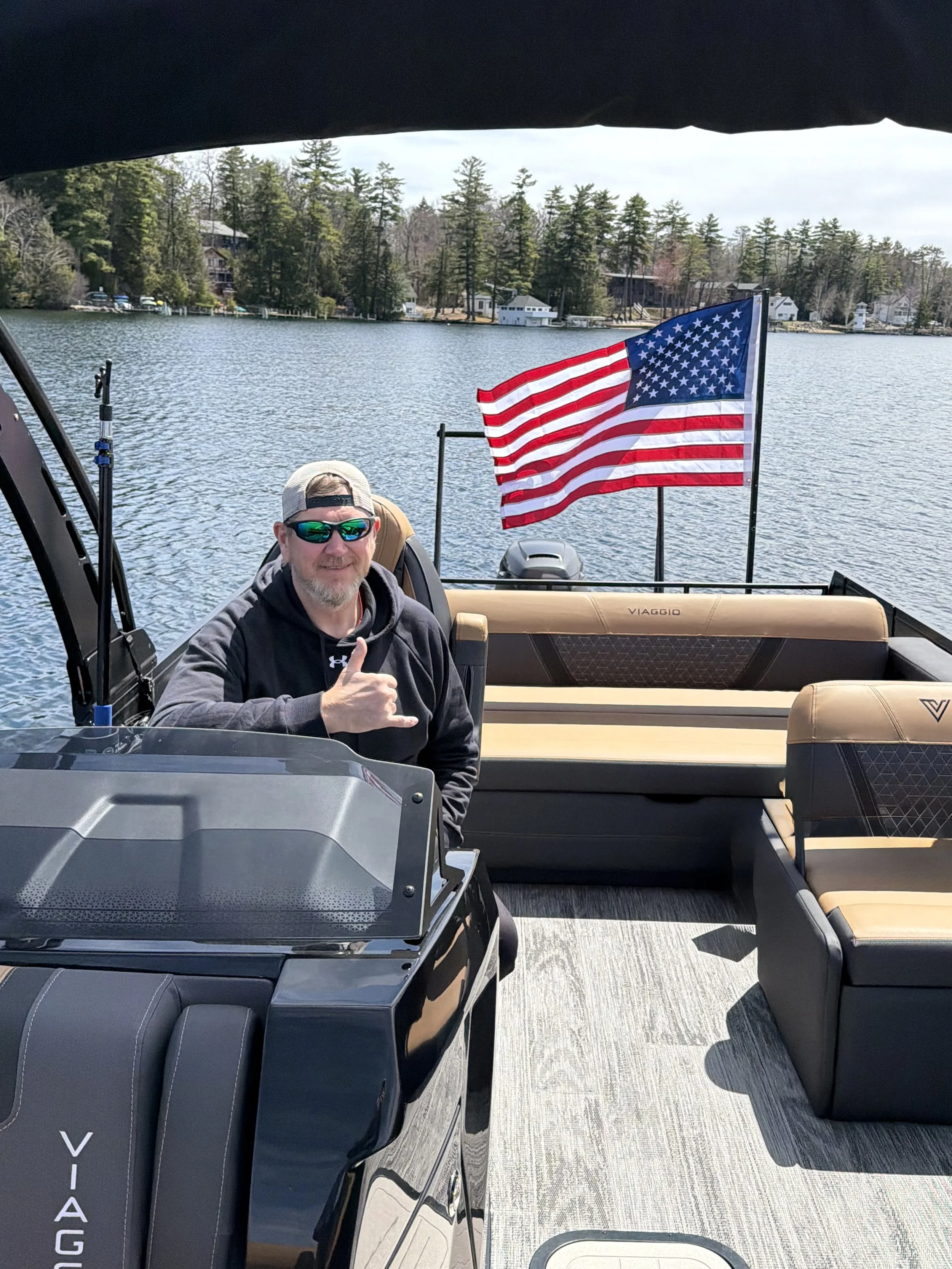 A man wearing sunglasses and a baseball cap sitting on a boat with American flag in the background. Lake and trees visible across the water.