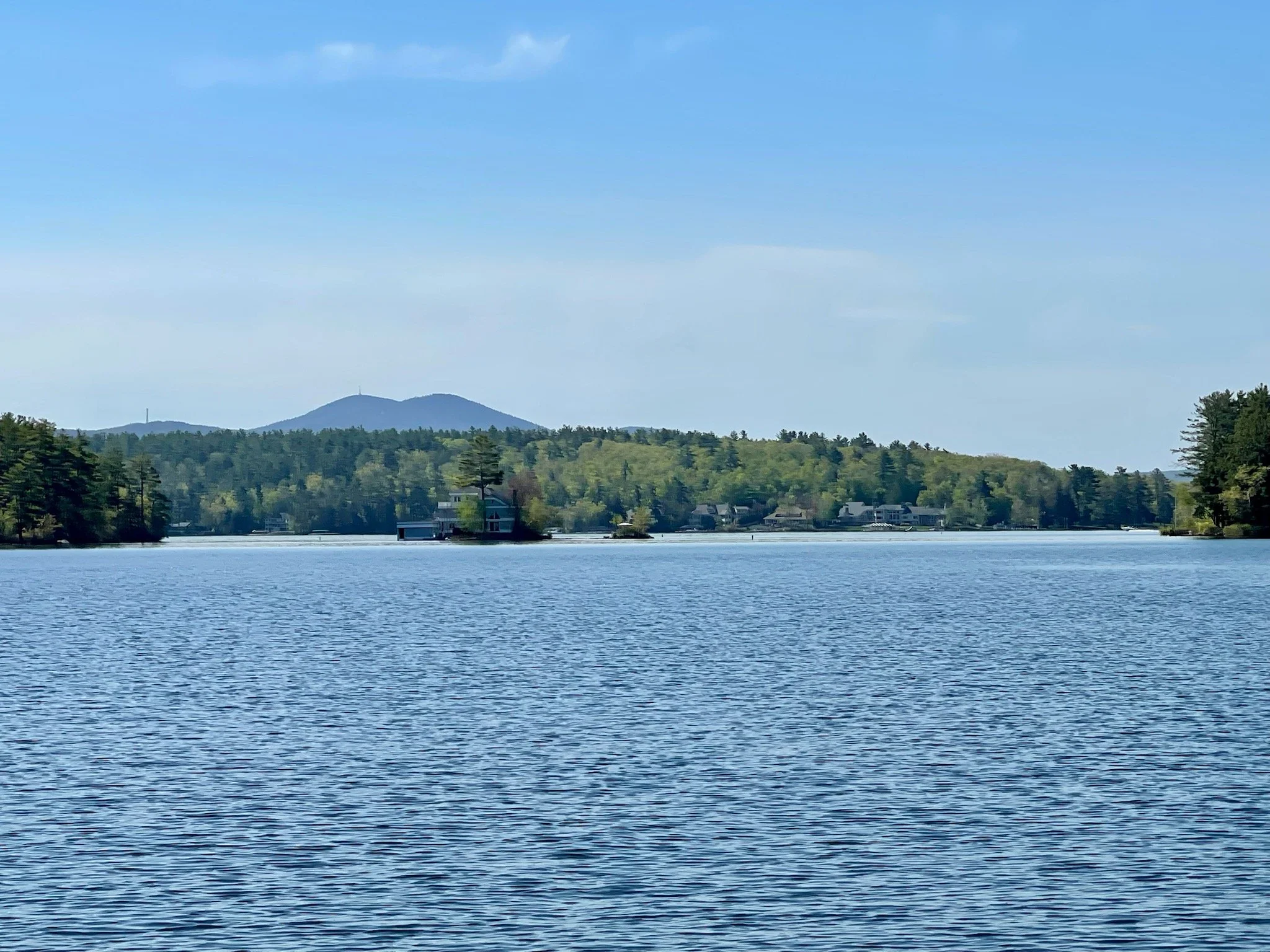 View of a lake with calm blue water, surrounded by green trees and houses, with mountains in the distance under a partly cloudy sky.