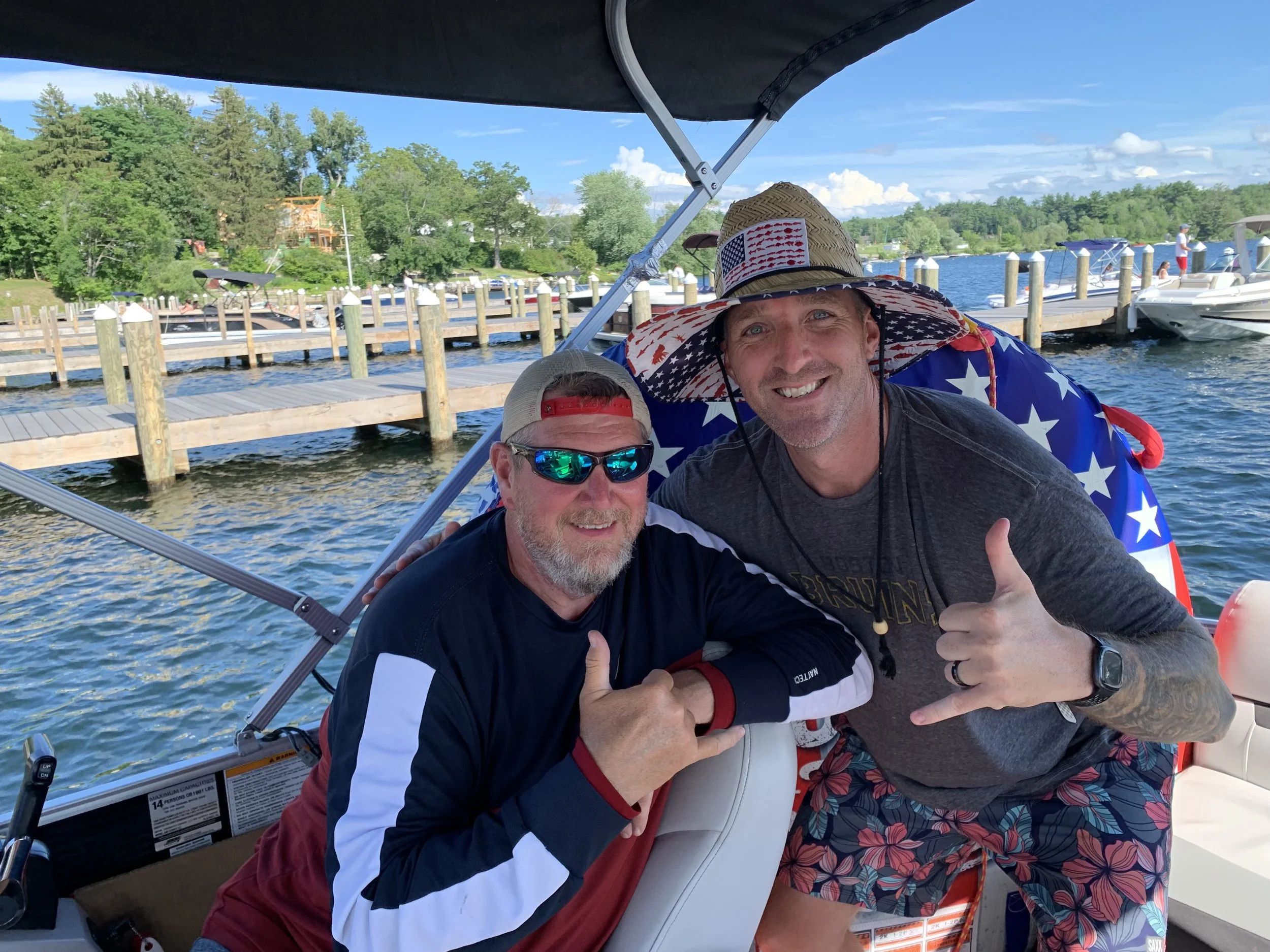 Two men are on a boat on a lake, smiling, giving thumbs up, and dressed in casual summer clothing. One wears a patriotic sun hat, and the other has sunglasses and a baseball cap. The background features a dock, boats, and lush green trees under a par