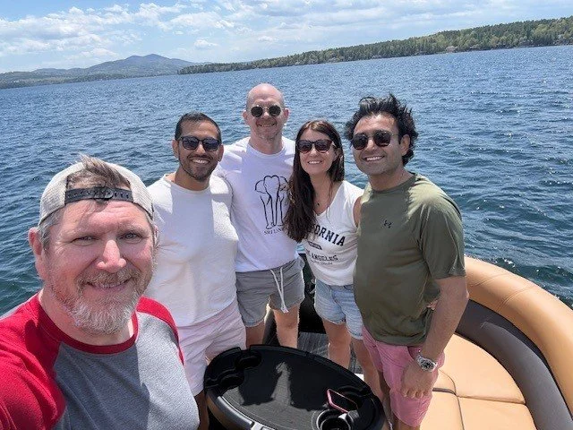 Group of five friends taking a selfie on a boat on a lake, with blue water, trees, and hills in the background, sunny weather.