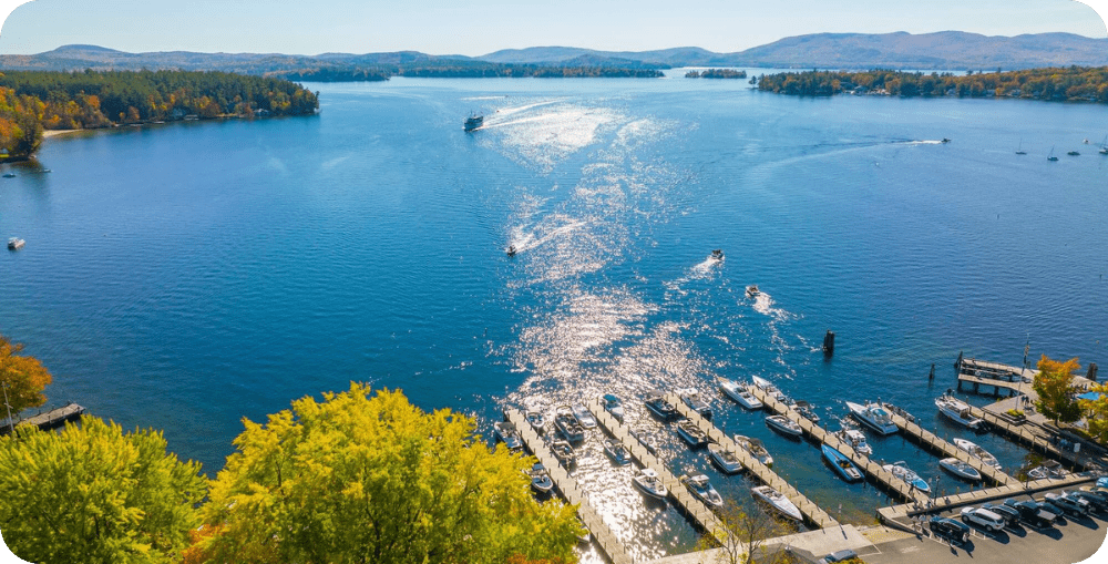 An expansive aerial view of Wolfeboro Bay on Lake Winnipesaukee, showing the town's historic waterfront, public docks, and a variety of boats on the sparkling blue water under a bright summer sky.