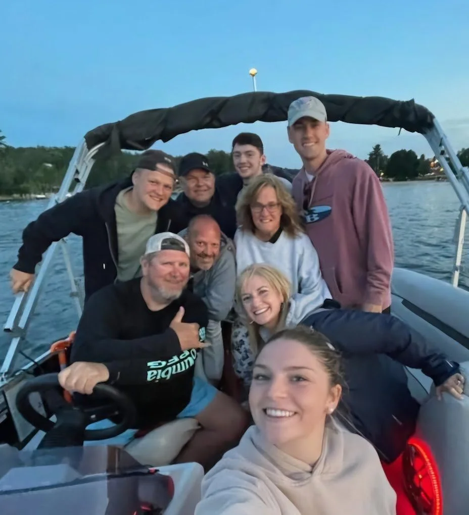 A group of nine people on a boat, smiling and posing for a selfie on a lake during sunset.