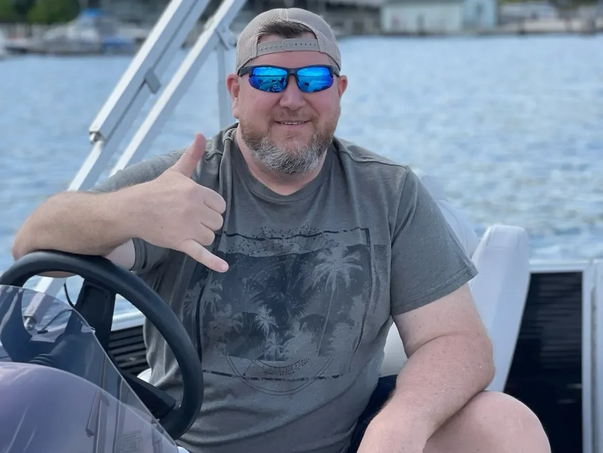 A bearded man wearing sunglasses, a baseball cap backwards, and a casual t-shirt is sitting on a boat near a body of water, smiling and giving a thumbs-up.
