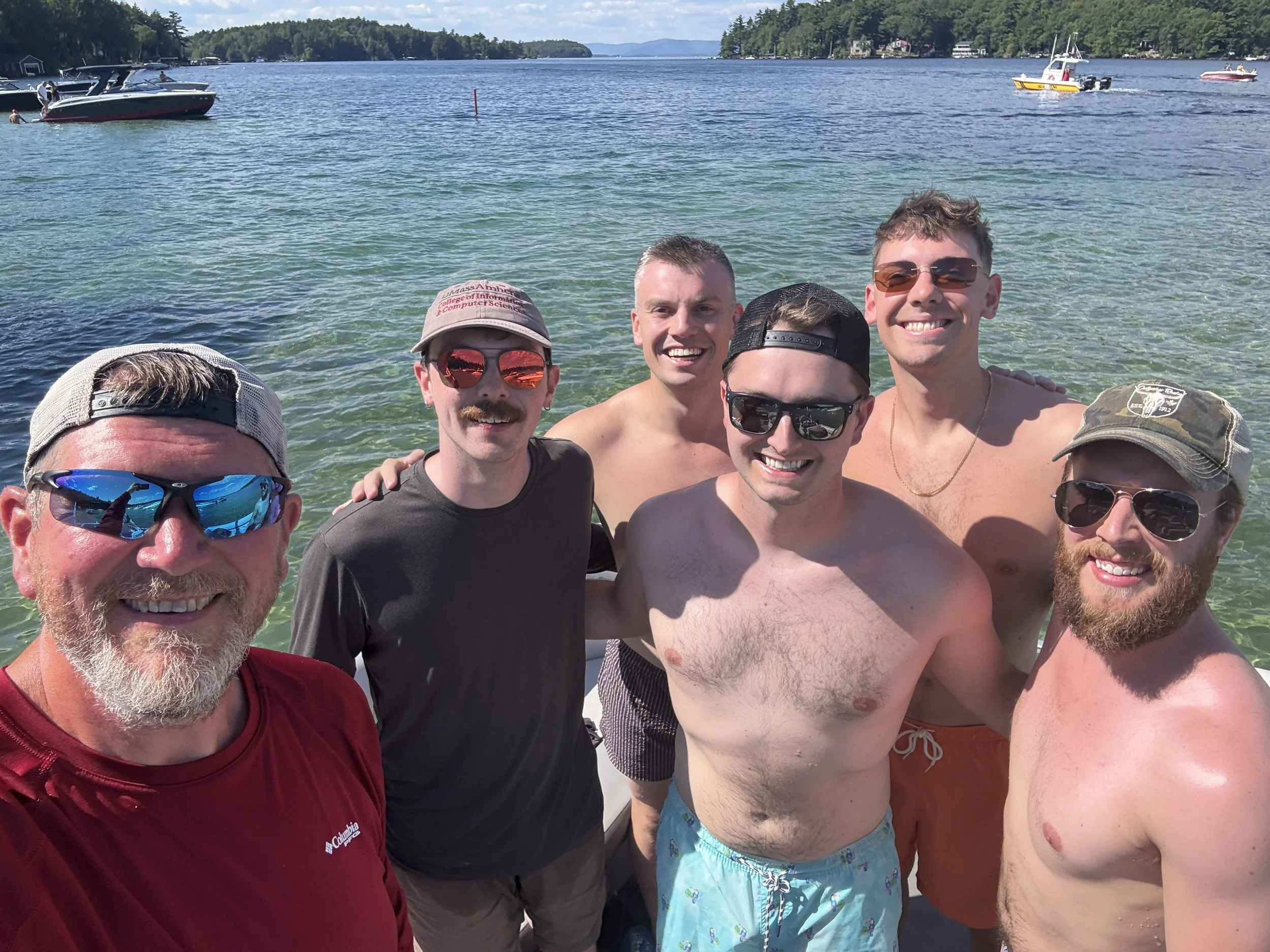 Group of men smiling and taking a selfie on a boat during sunny day on a lake, with multiple boats and trees in the background.