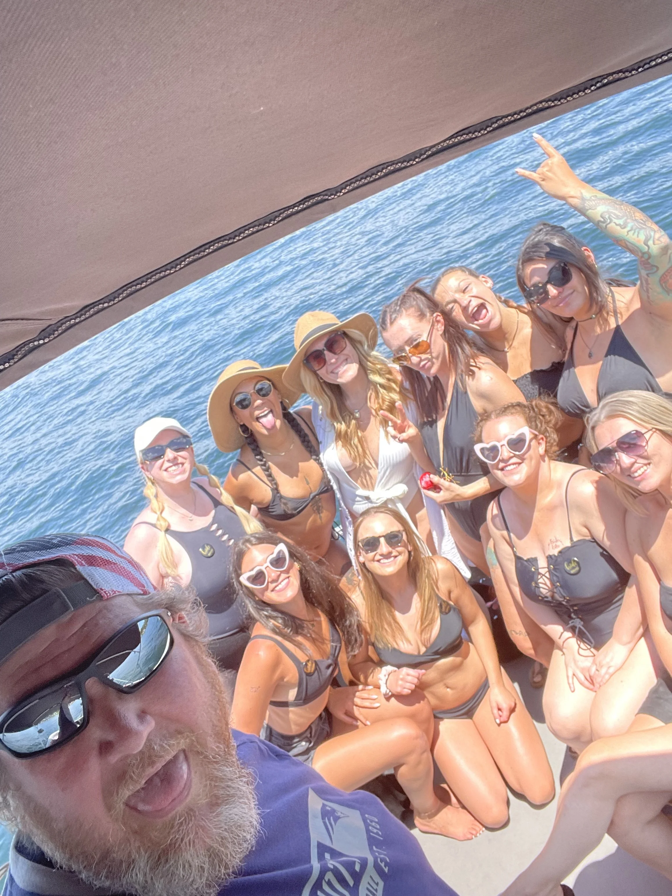Smiling group of women in swimsuits and sunglasses on a boat at the beach, with blue water in the background, taking a selfie with a bearded man in sunglasses in the foreground.